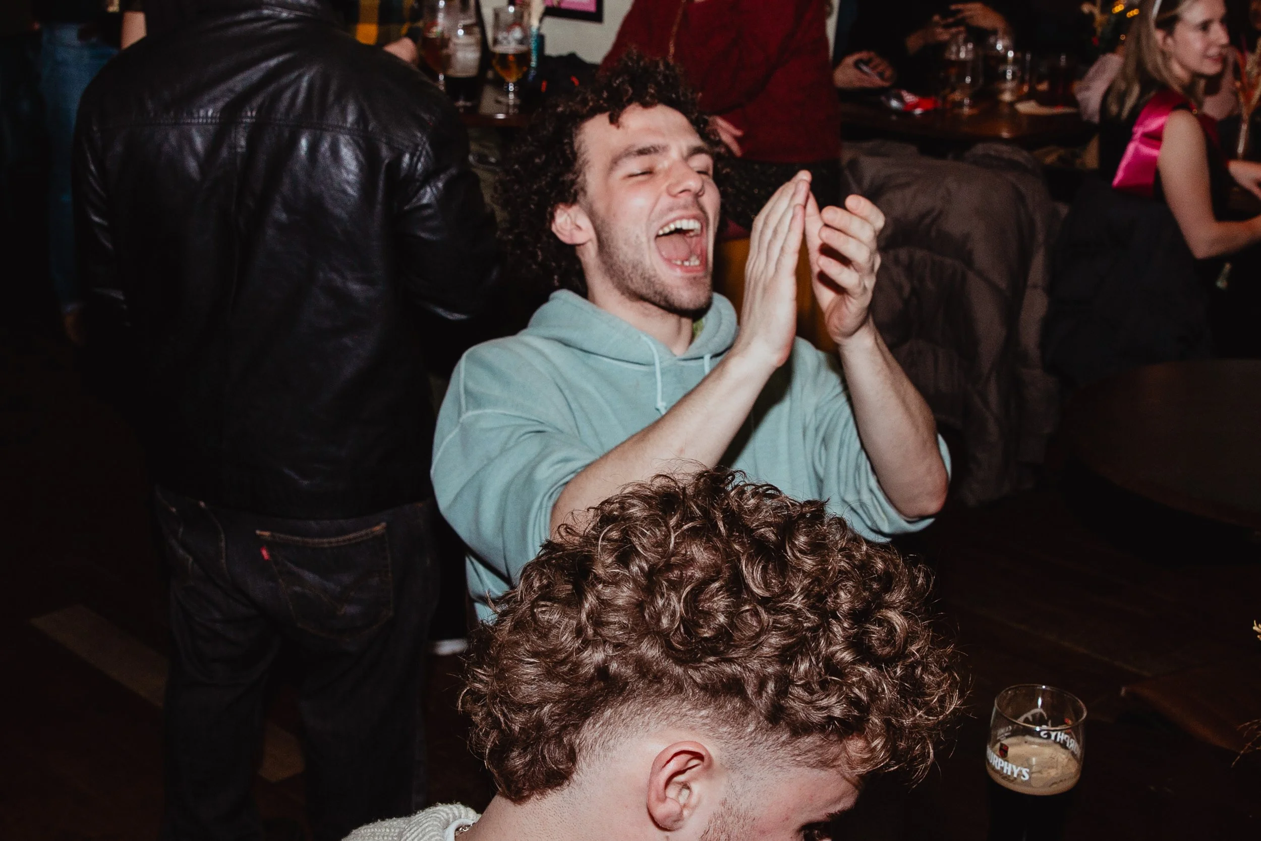 A man with curly hair claps and laughs at a lively gathering in a bar or pub.