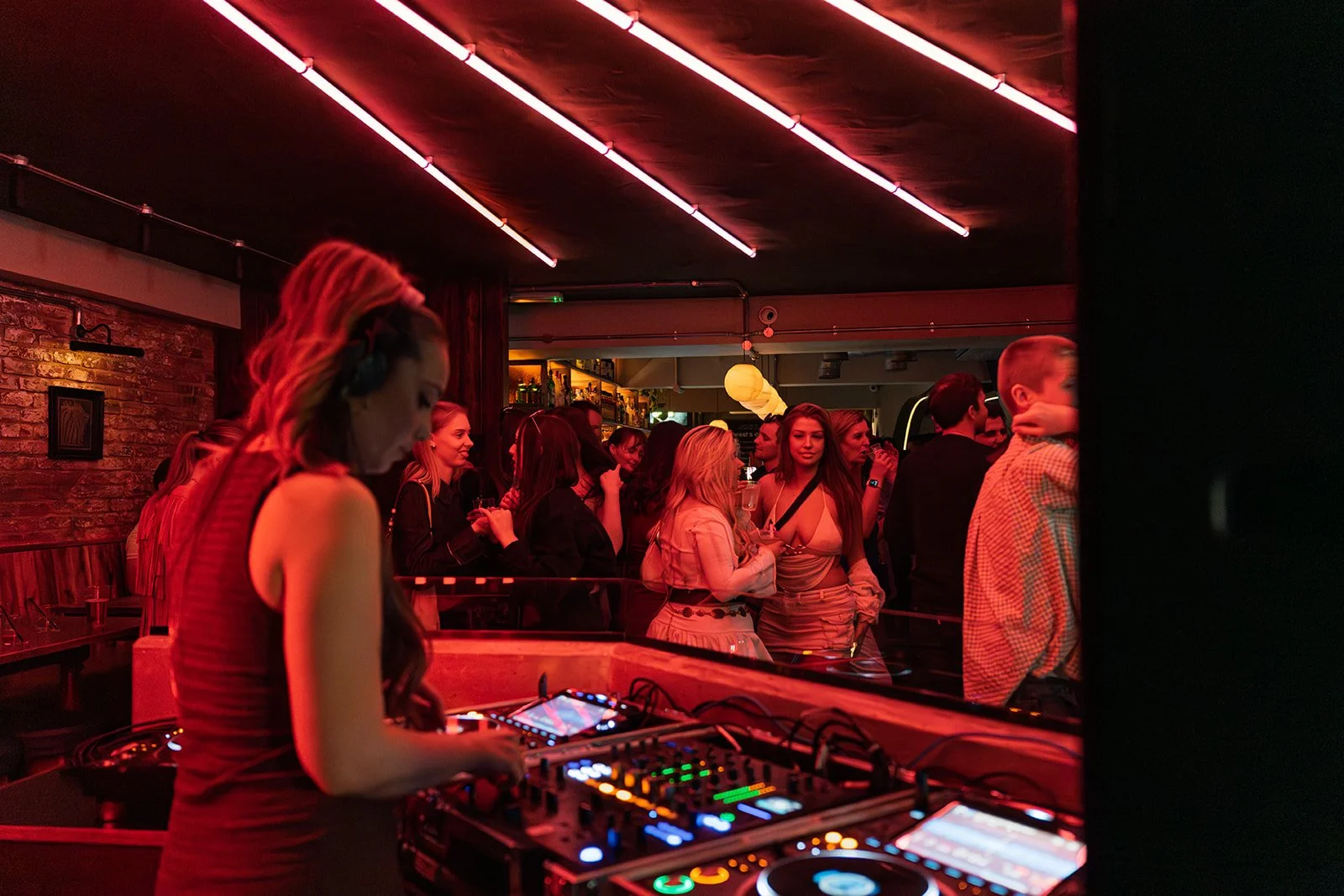 A DJ performing at a nightclub with a crowd of people dancing and socializing in the background, illuminated by red and pink neon lights.