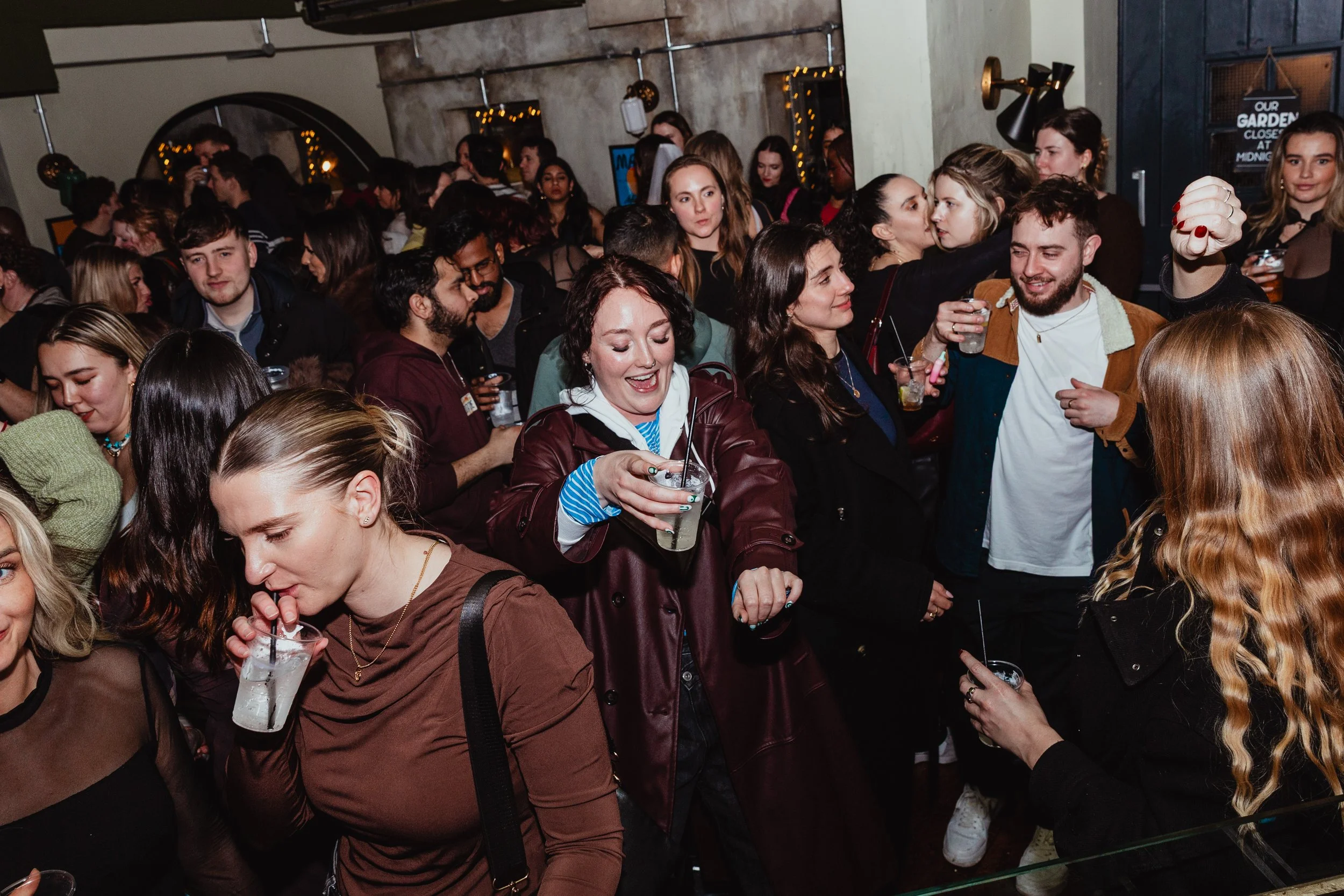 Crowd of people enjoying a lively party or social gathering indoors, with some holding drinks, smiling, and engaging in conversation.