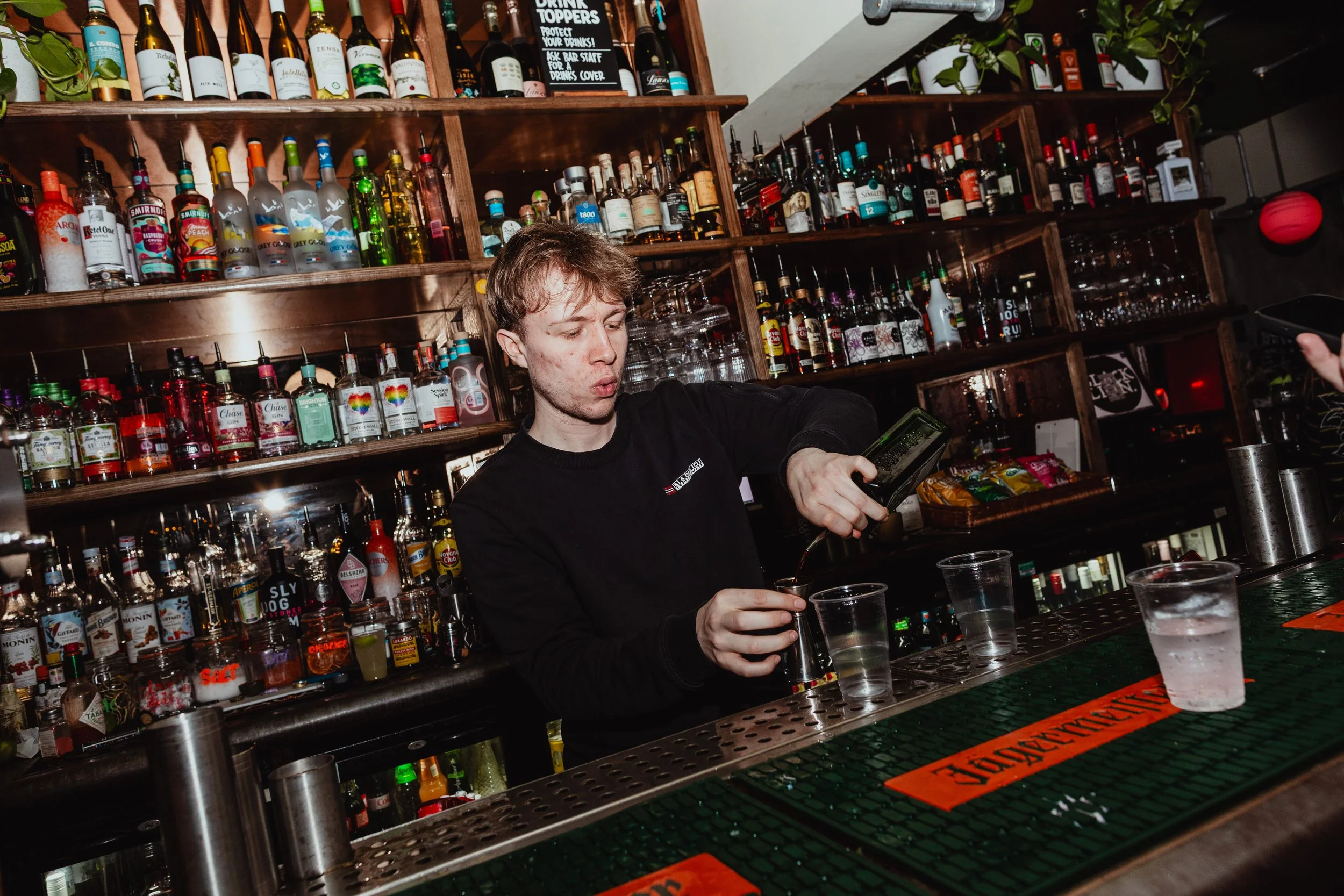 A bartender pouring a drink at a bar with shelves of various alcohol bottles behind him.