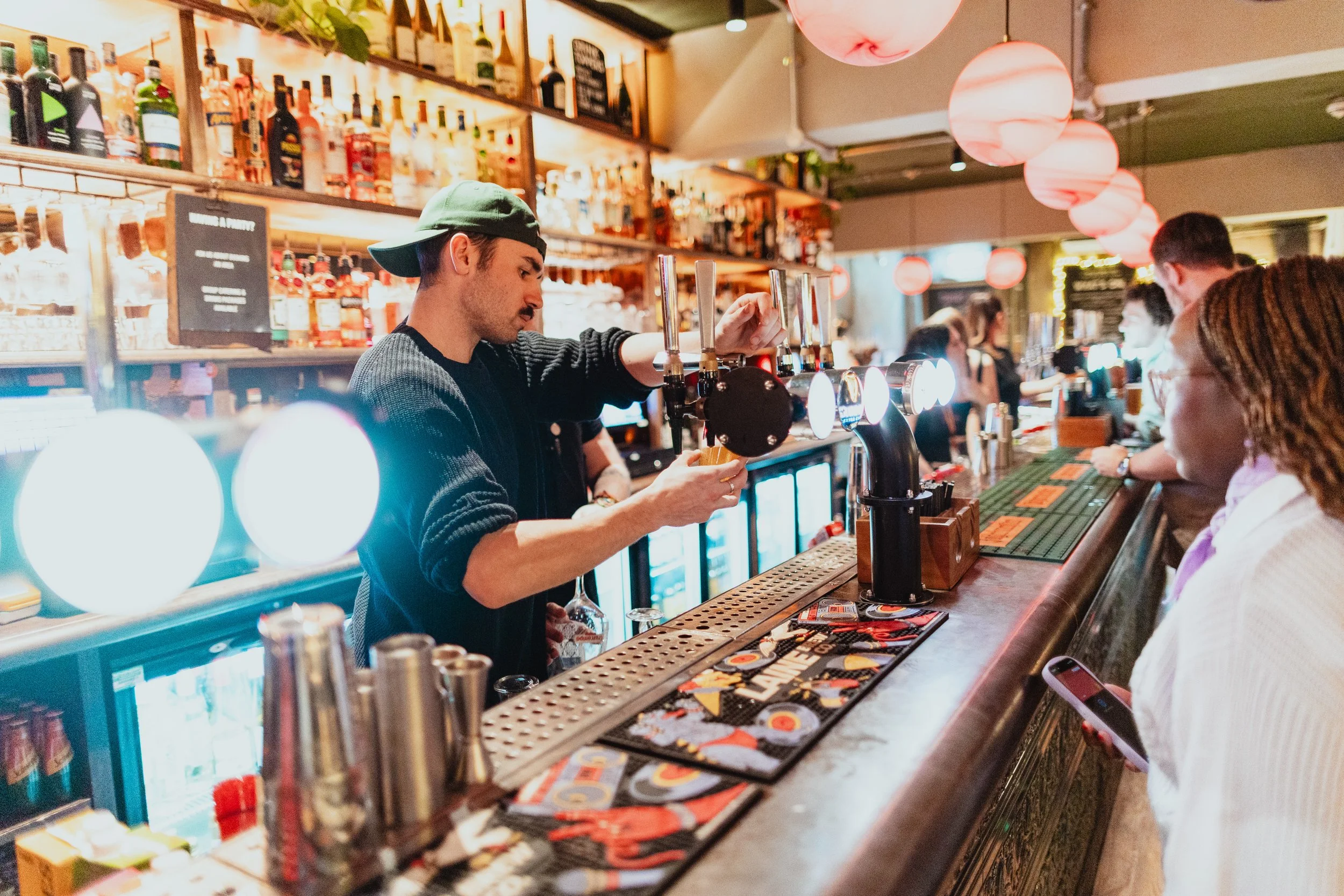 Bartender pouring draft beer for a customer at a bar with bottles of alcohol on shelves behind. Several patrons seated along the bar, some holding drinks or looking at their phones, with pink globe lights overhead.