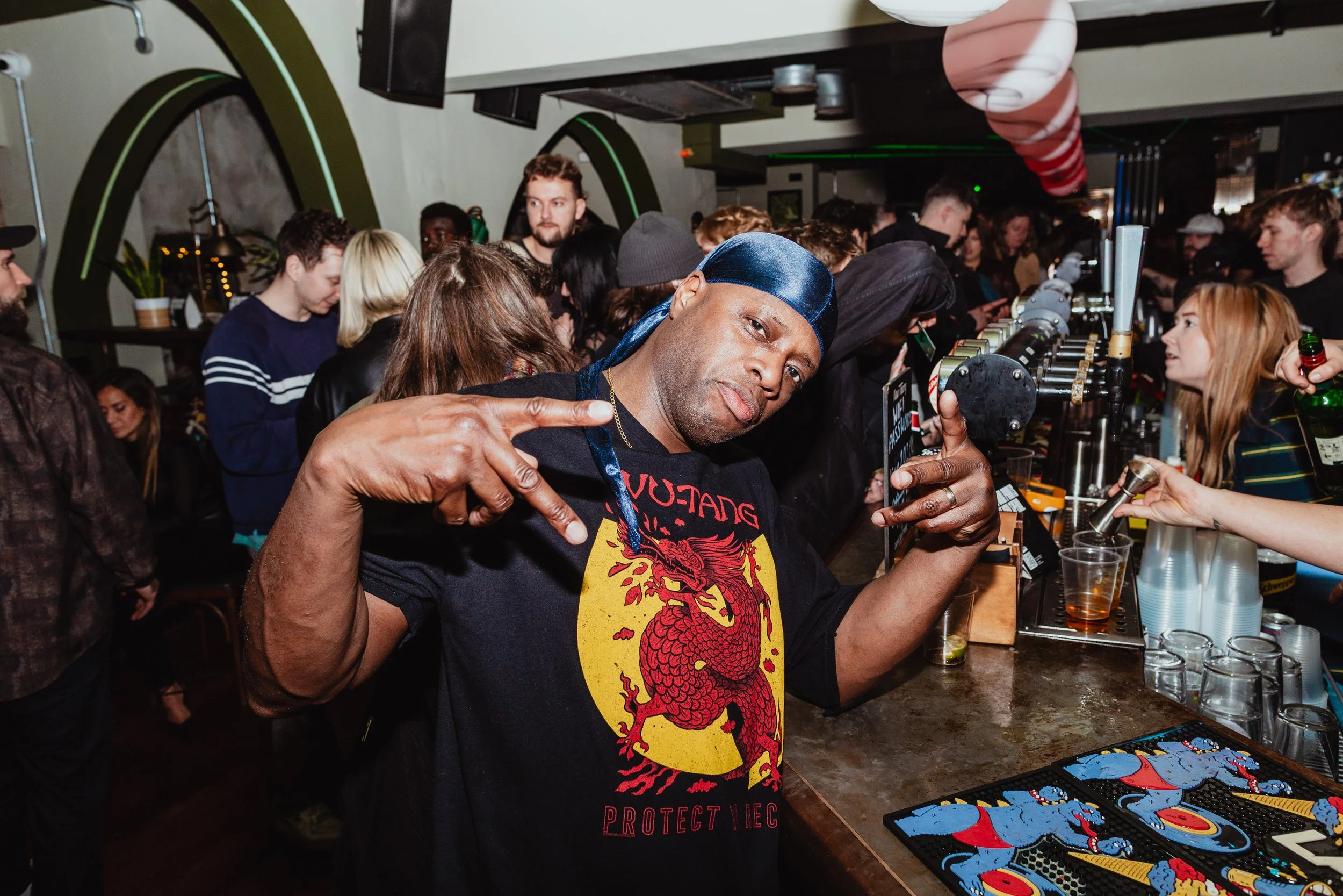 A man at a crowded bar or nightclub posing with peace signs, wearing a black t-shirt with a red and yellow dragon graphic and a blue headscarf, with people and bar equipment in the background.