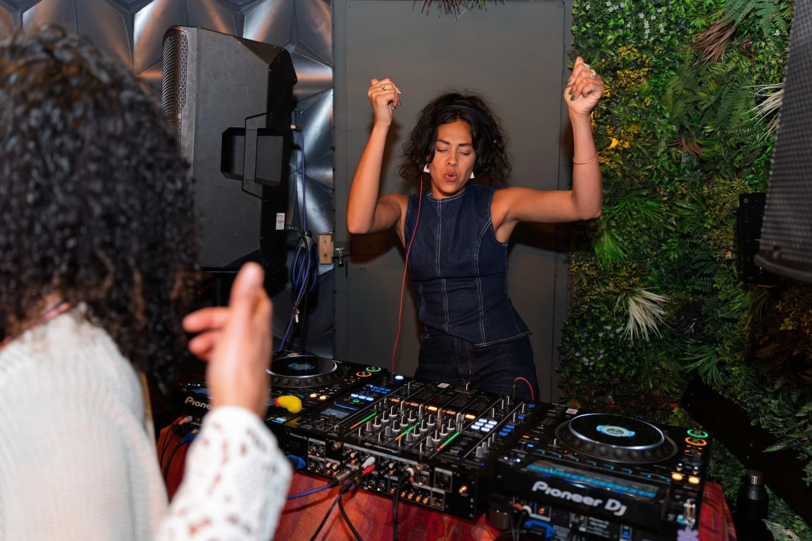 A woman with curly hair dancing with her eyes closed at a DJ booth, wearing headphones, in a space with a green leafy wall, while another person in the foreground watches and claps.
