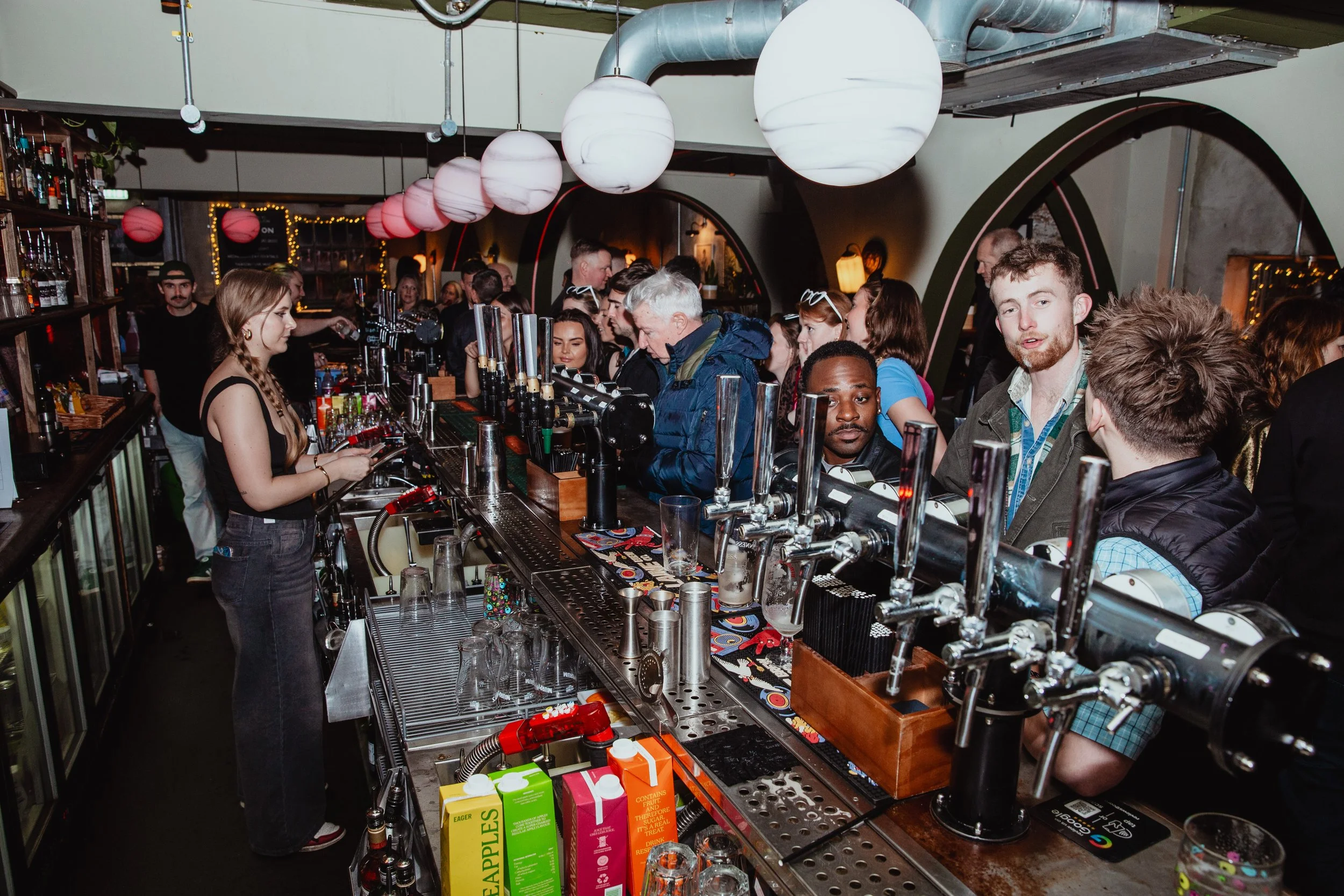 Crowded bar with people ordering drinks, bartender preparing beverages, lit decorative lanterns hanging from ceiling.