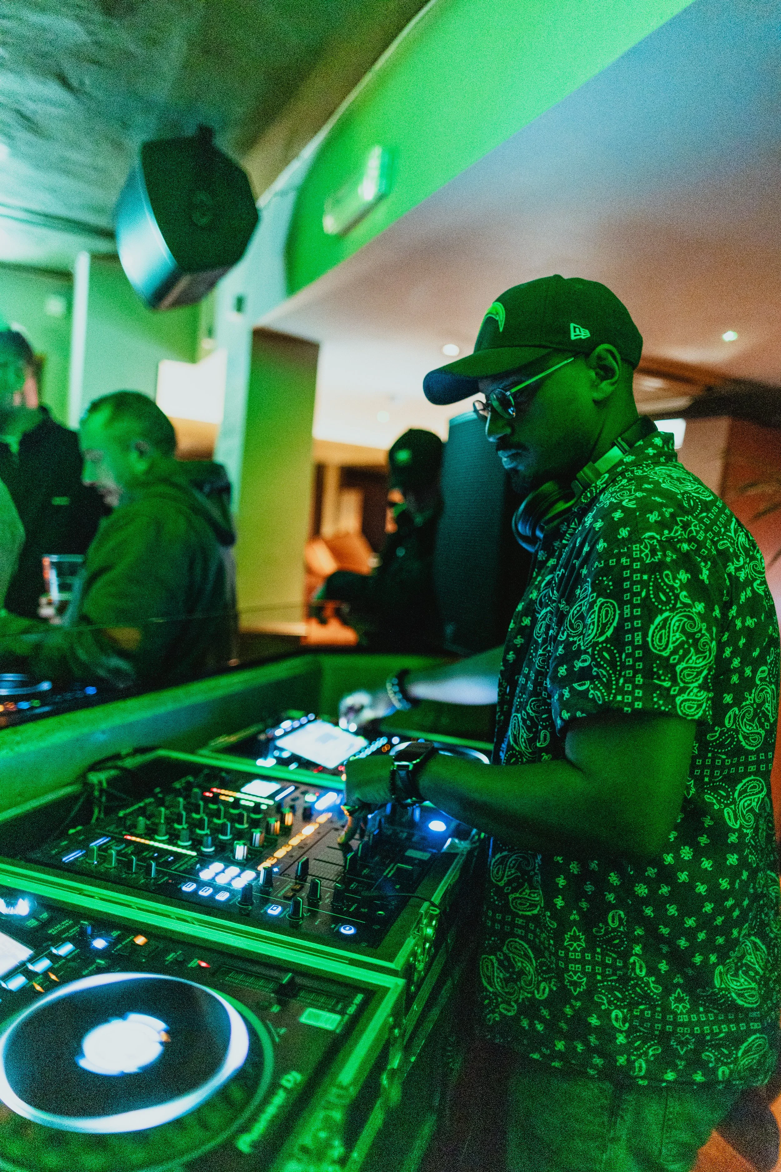 A DJ wearing a cap, glasses, and headphones rides a DJ controller at a party, with people dancing in the background.
