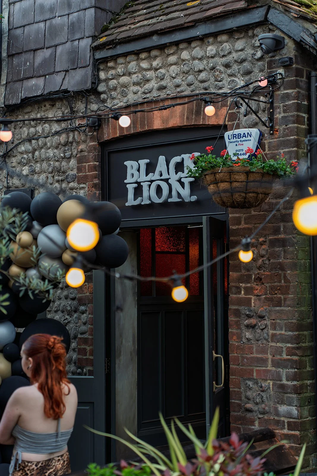 The exterior of a venue called 'Black Lion' with a black door, brick and stone wall, hanging flower basket, string lights, and decorative black, gold, and gray balloons.