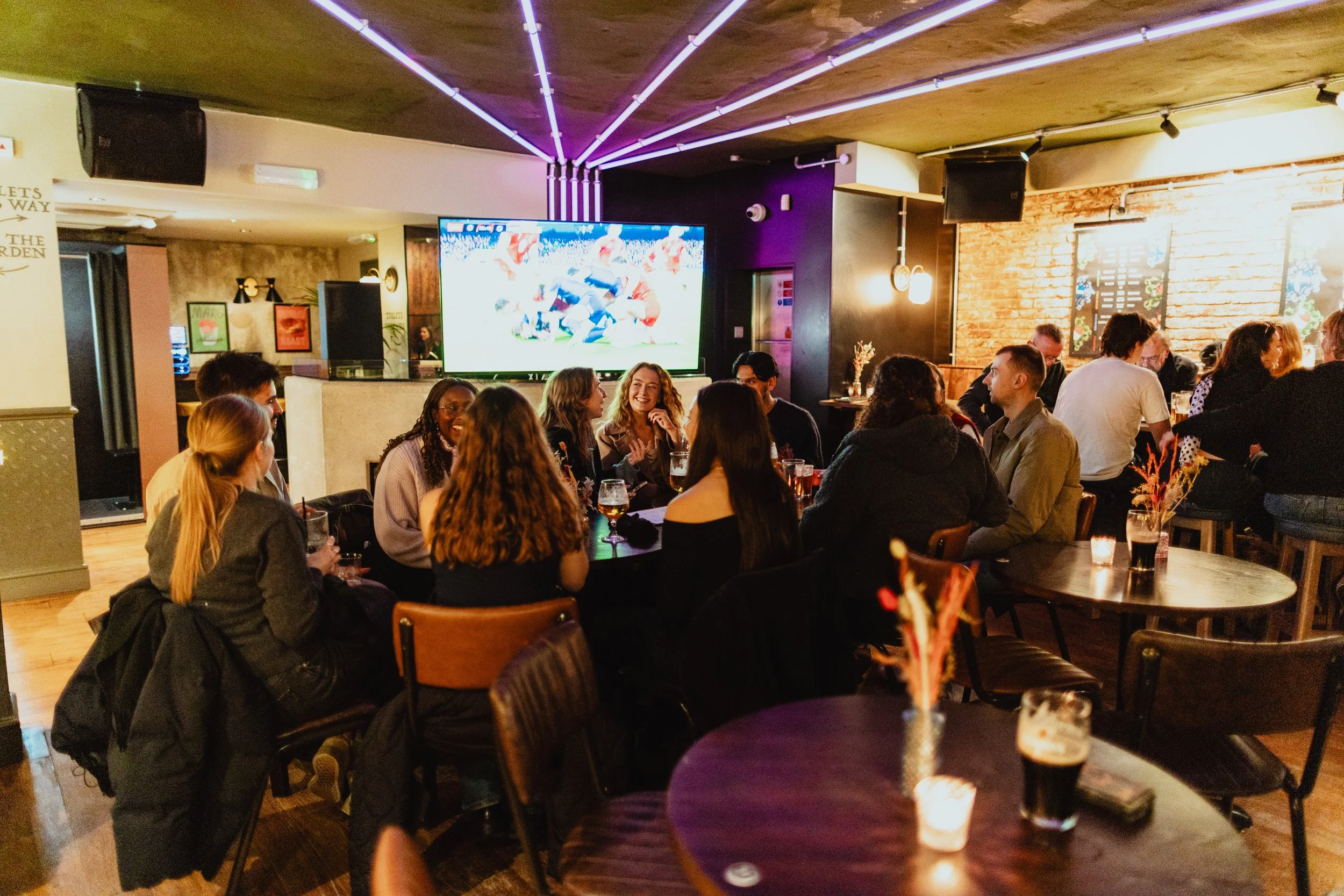 Group of friends socializing at a bar or restaurant, watching a sports game on a large TV, with drinks on the tables and dim, cozy lighting.