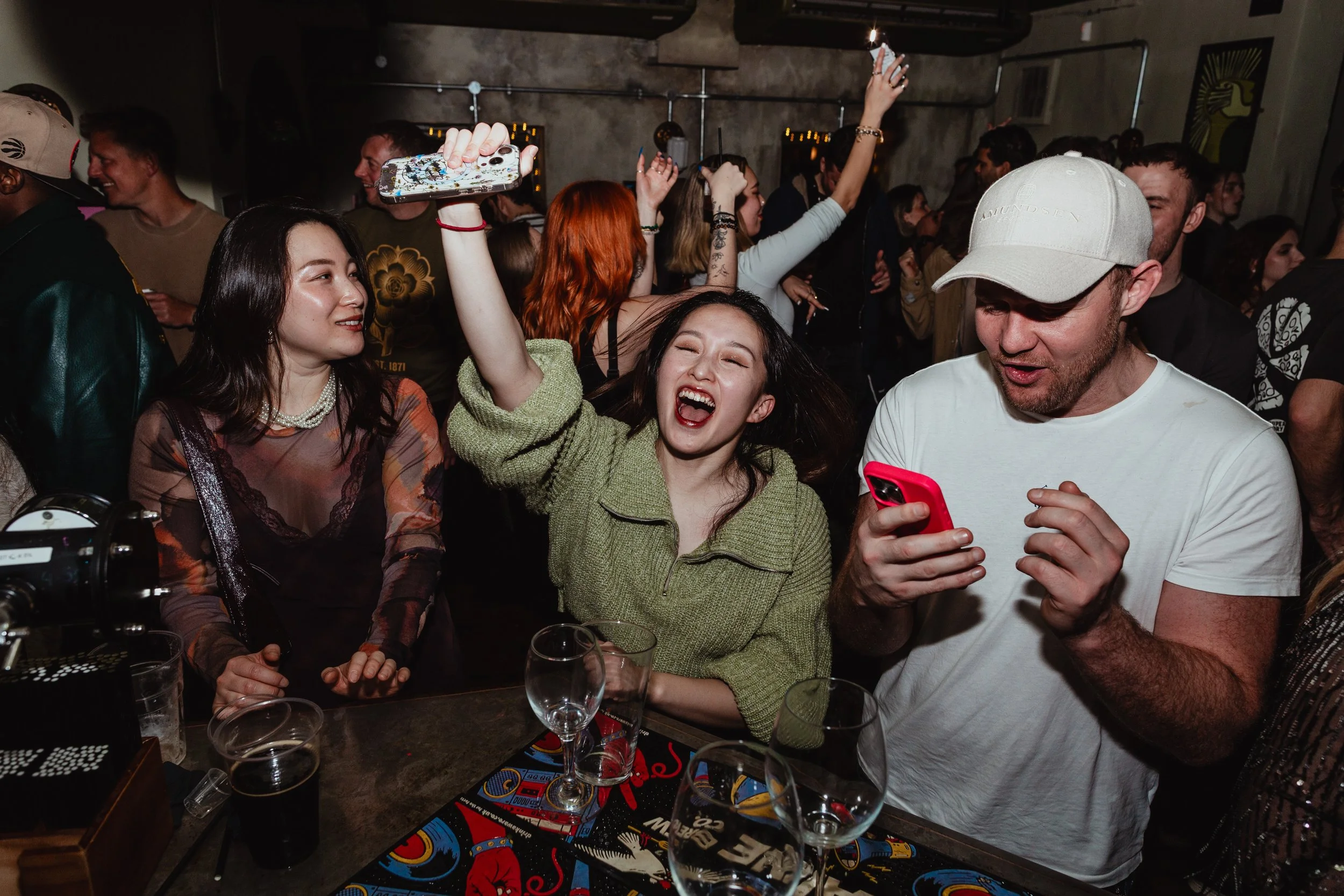 Group of friends enjoying a party at a bar, with one woman dancing and smiling while others look at their phones.