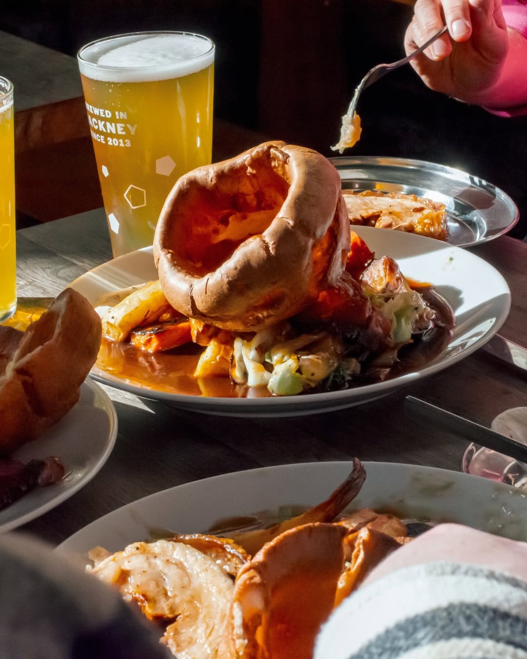 A meal with a large Yorkshire pudding on a plate, served with vegetables and gravy. There are glasses of beer in the background, and a person is holding a fork with food. The setting appears to be a restaurant.
