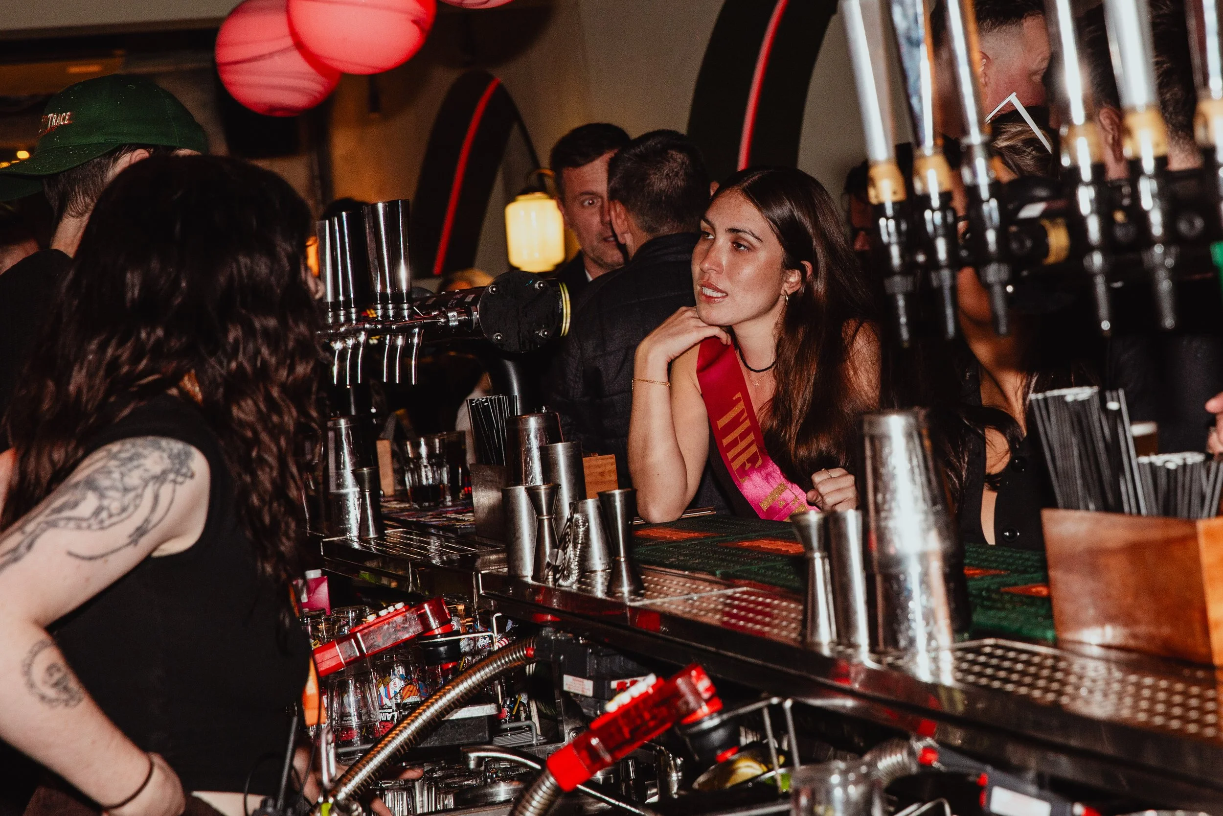 A woman with long dark hair, wearing a pink sash, sitting at a bar counter talking to a bartender with tattooed arm, and other people in the background at a lively bar or club.