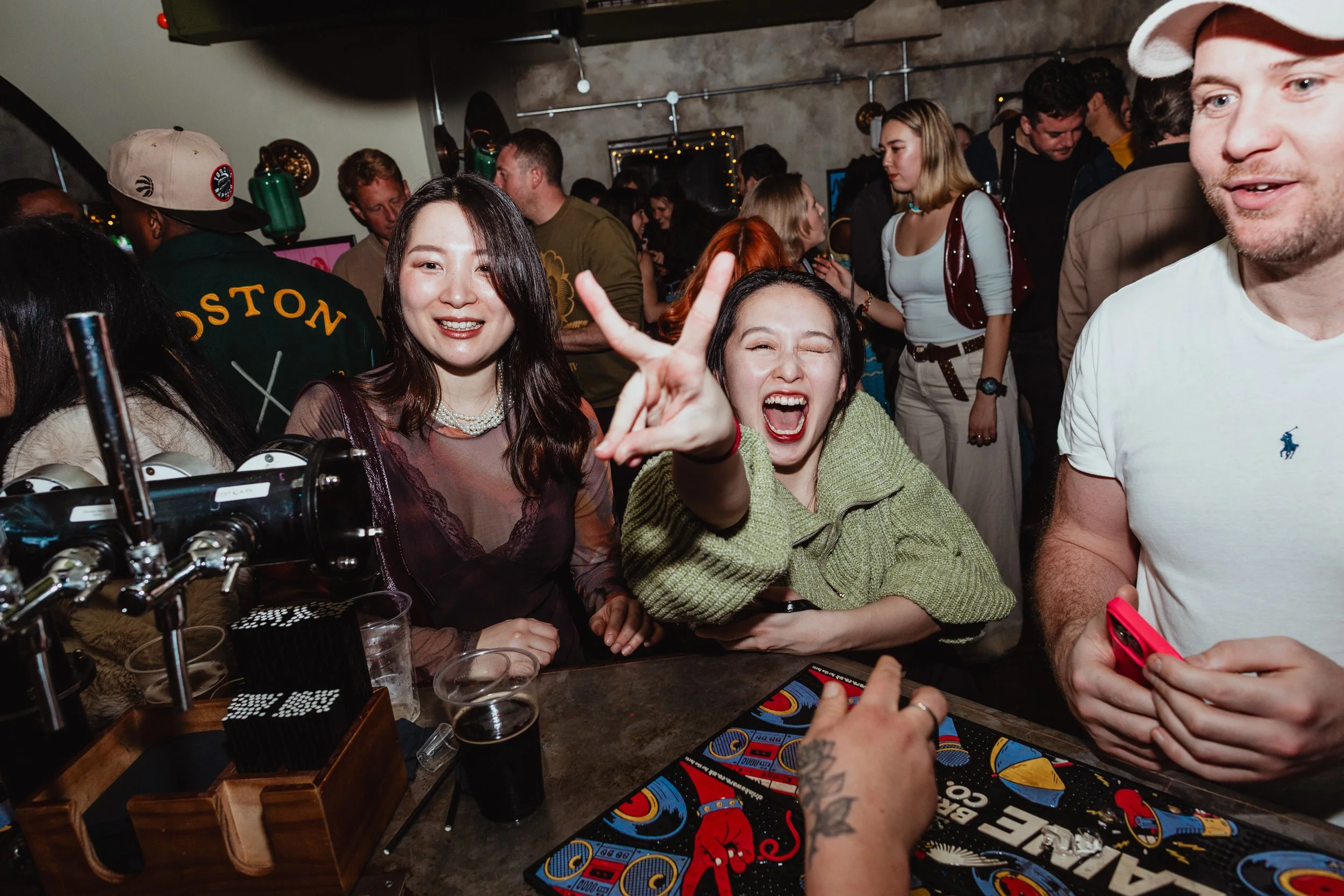People enjoying themselves at a crowded bar or party. Two women in the foreground are cheerful, one making a peace sign, with drinks and game cards on the table. In the background, people are socializing in a lively atmosphere.