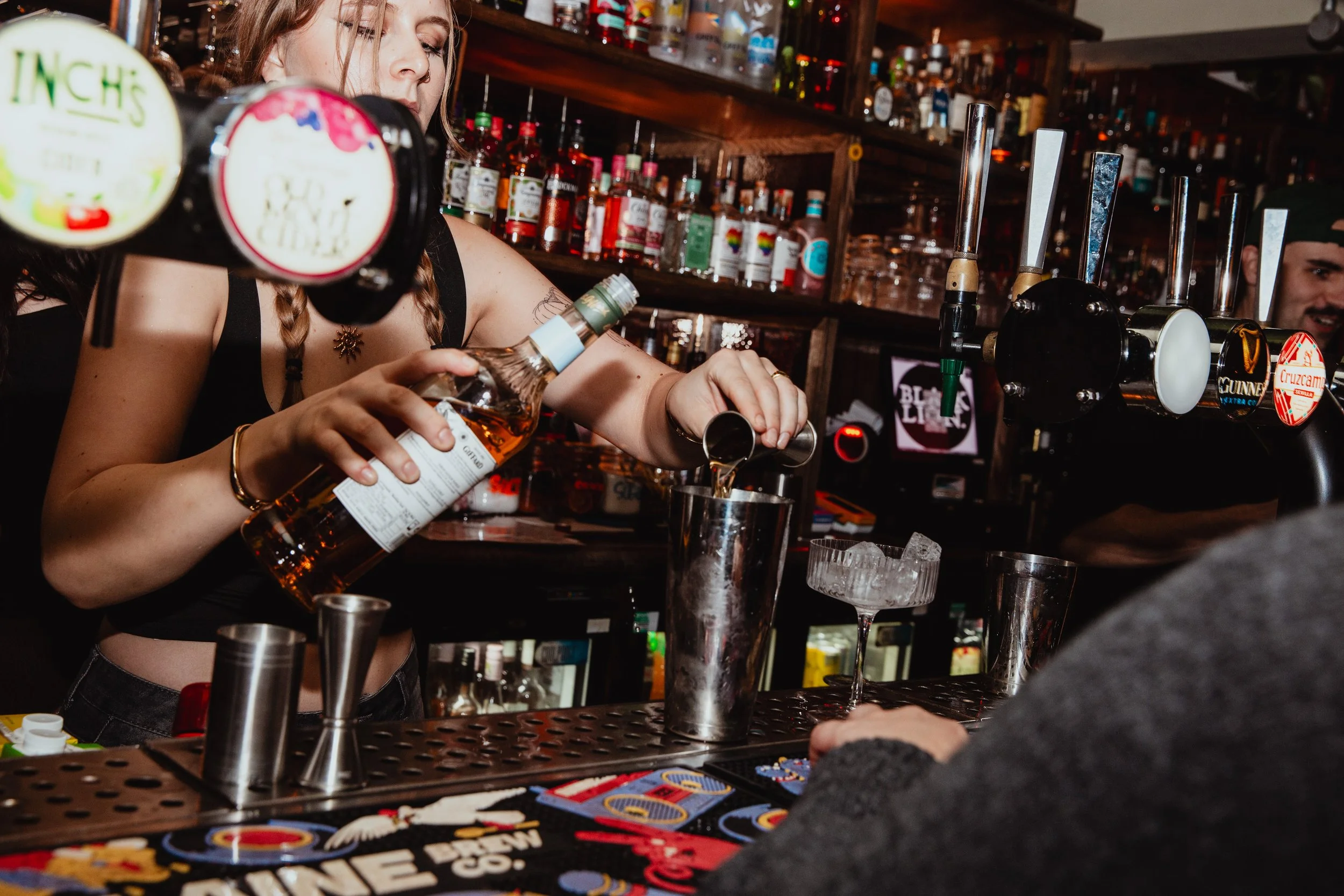 A bartender pouring a drink into a metal shaker behind a bar with liquor bottles and taps in the background.