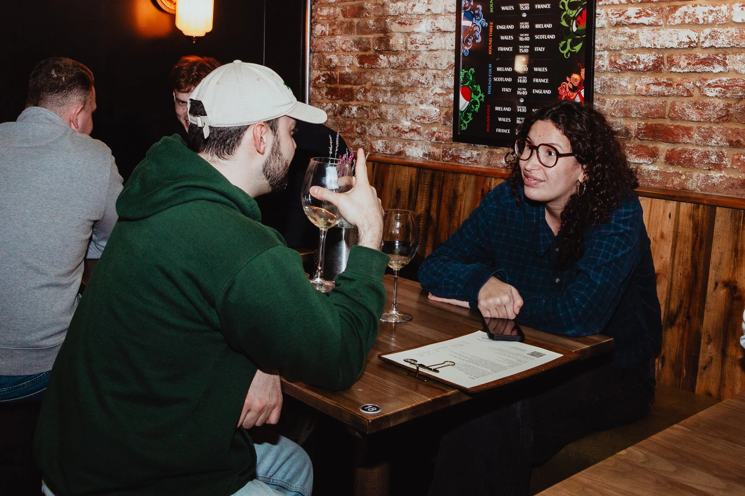 Two people sitting at a wooden table in a bar, engaged in conversation. The man wearing a green hoodie and white cap, holding a glass of white wine, and the woman with curly hair and glasses, with a menu and smartphone on the table. There are other p