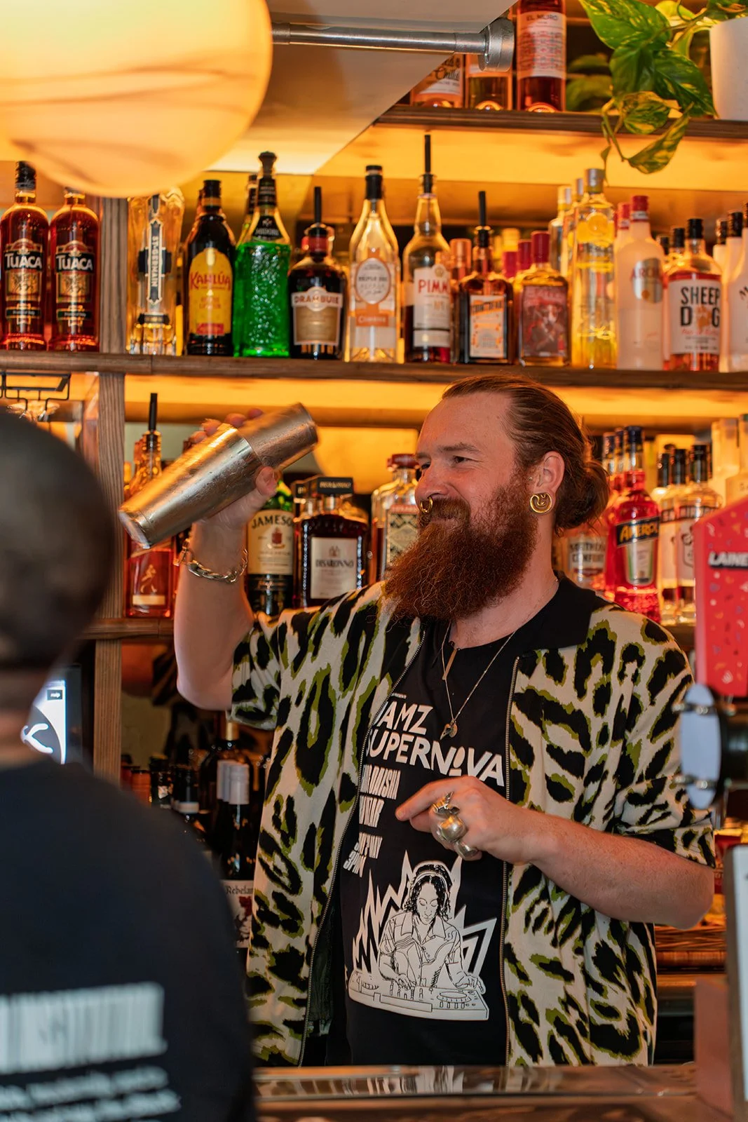A bartender with a beard, piercings, and a leopard print shirt shaking a cocktail shaker behind a bar. The bar shelf is stocked with various bottles of liquor.
