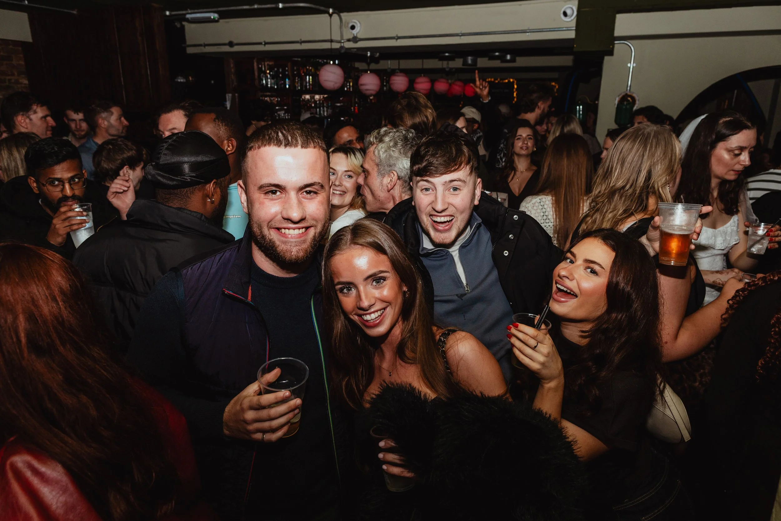 A group of four young people smiling and posing in a crowded bar or party setting with drinks in their hands, surrounded by other partygoers.