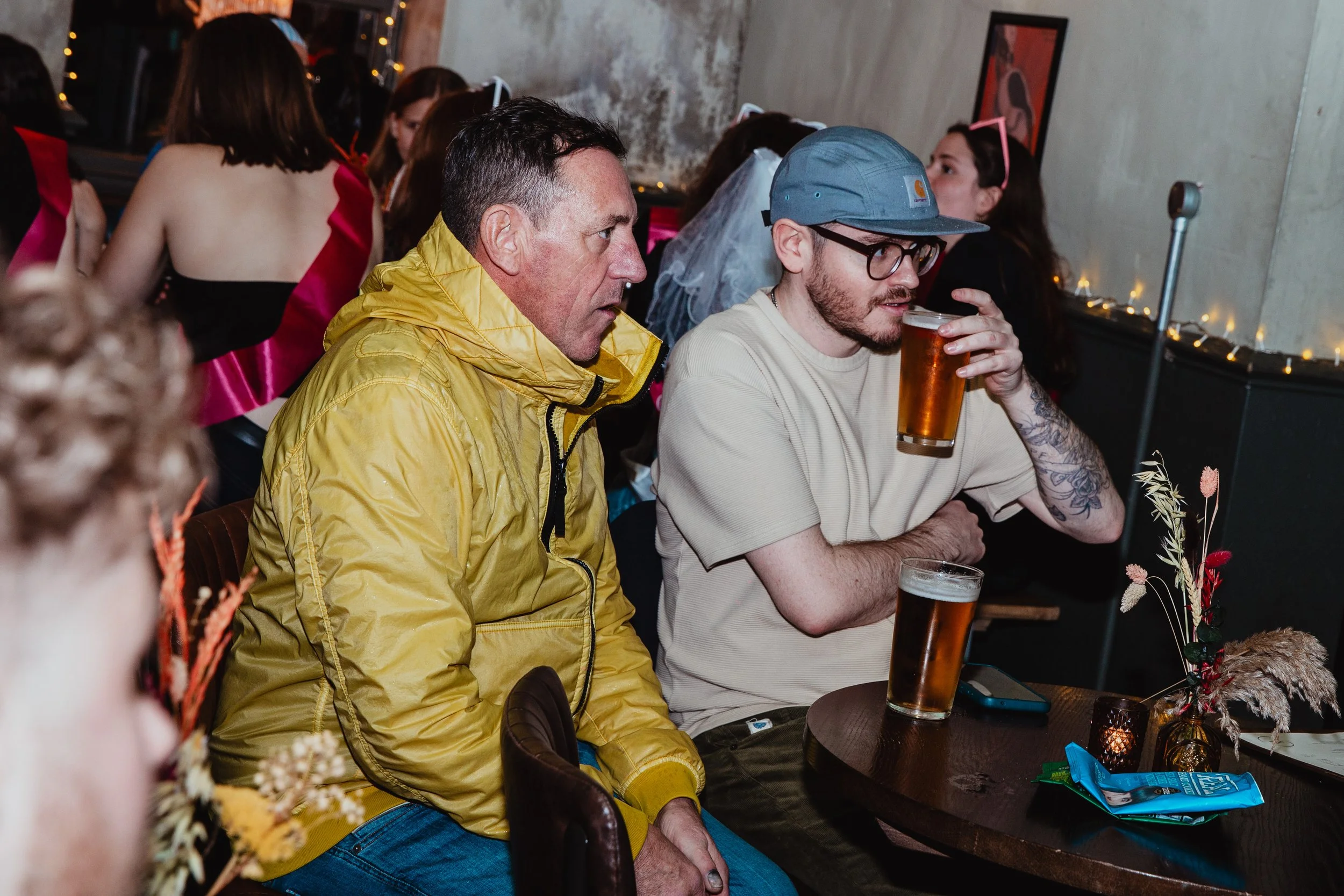 Two men sitting at a table in a dimly lit bar or restaurant, each holding a glass of beer. The man on the left is wearing a yellow windbreaker, while the man on the right is wearing a beige T-shirt, glasses, and a blue cap. In the background, there a