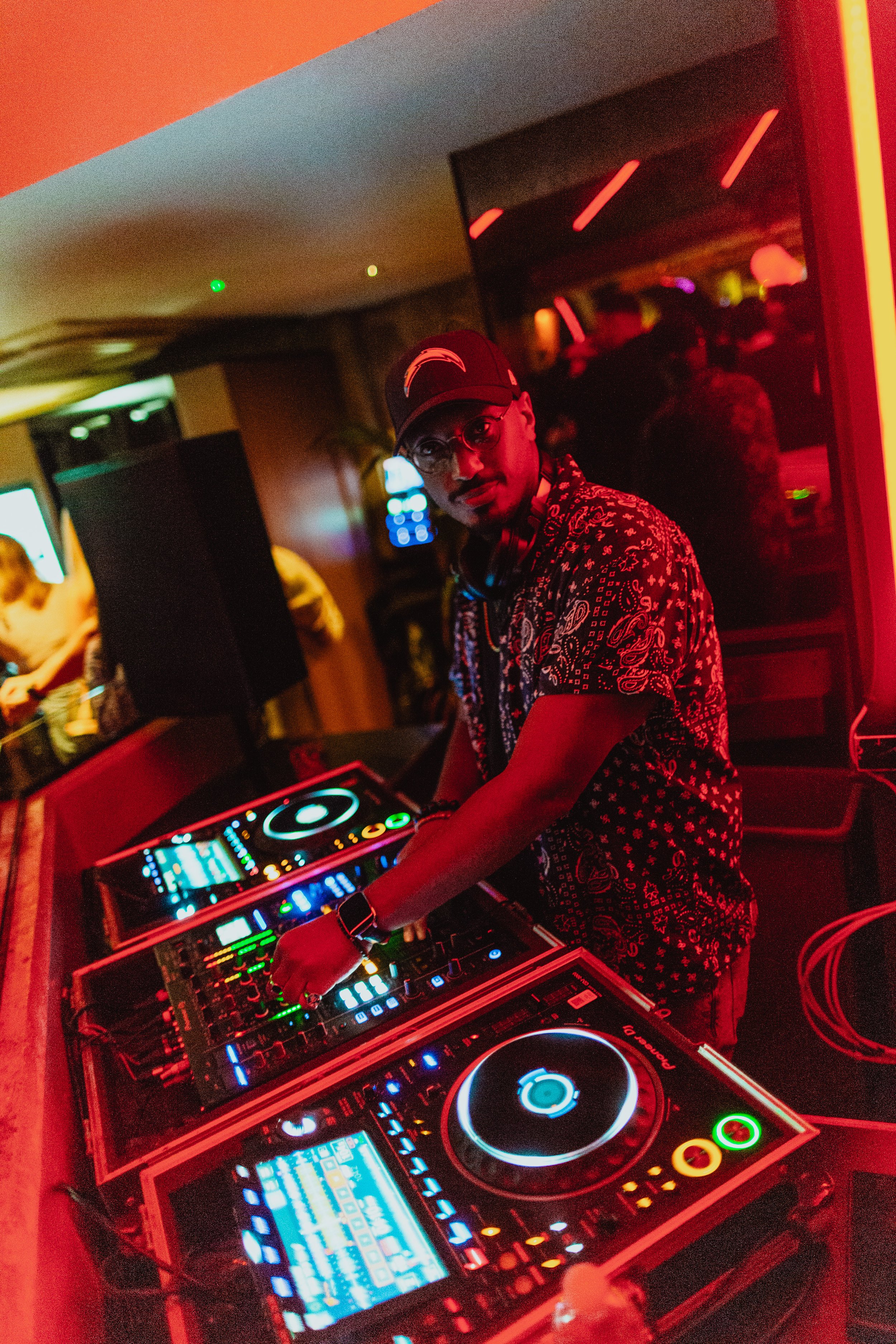 A DJ wearing glasses, a cap, and headphones around his neck, stands behind a DJ console in a dimly lit nightclub with red and orange neon lighting, and a crowd in the background.