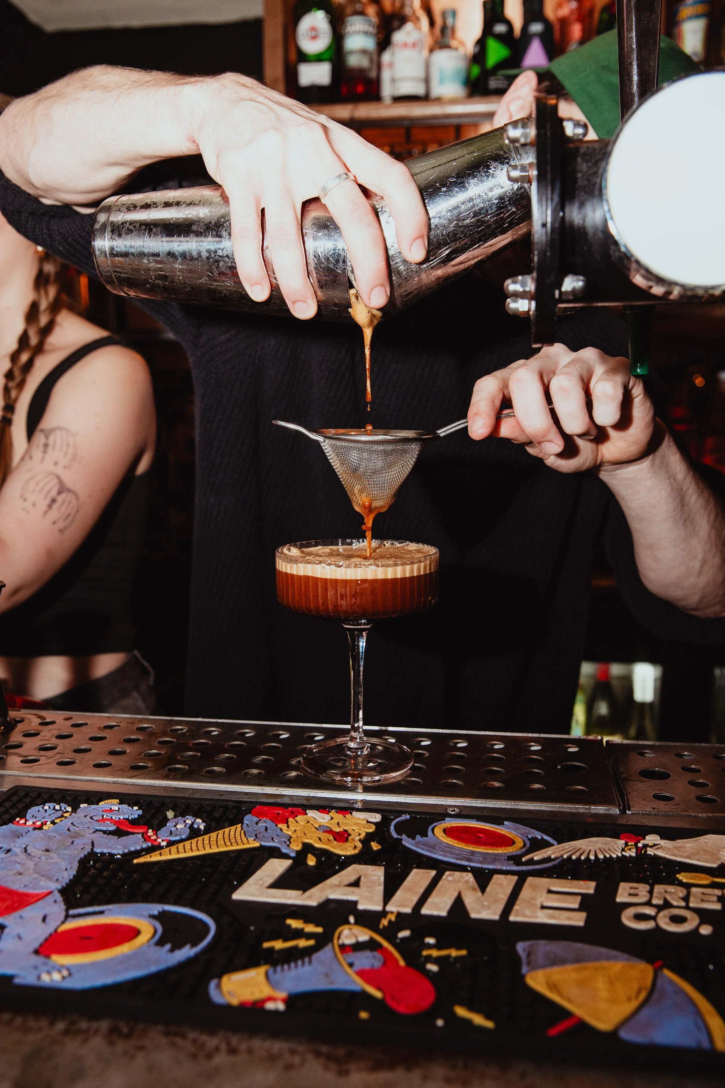 A bartender pours a cocktail through a fine mesh strainer into a coupe glass. The bartender is using a cocktail shaker, and the scene is set at a bar counter with bottles in the background.