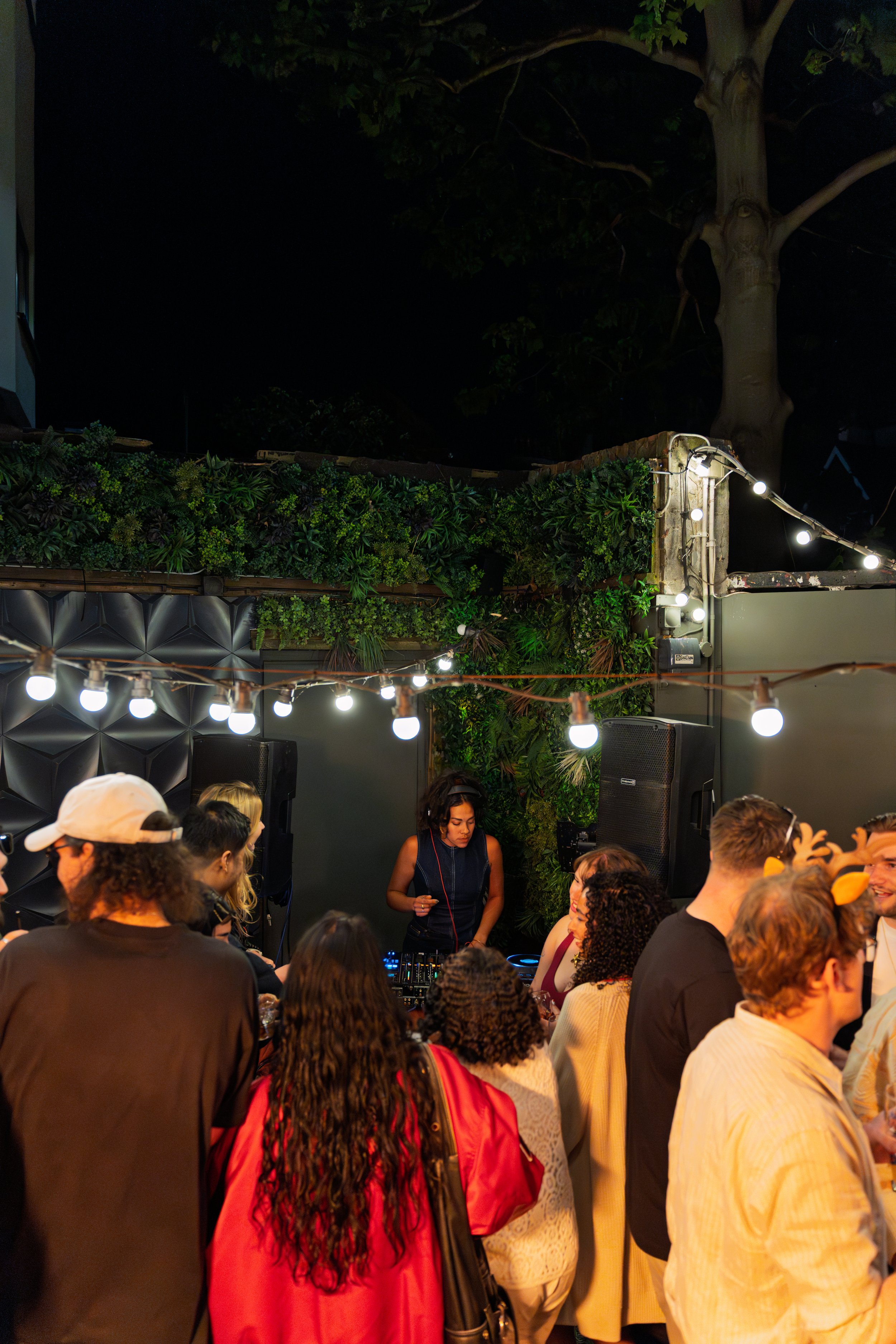 Nighttime gathering with a DJ playing music, surrounded by people socializing under string lights at an outdoor venue with greenery and a large tree in the background.