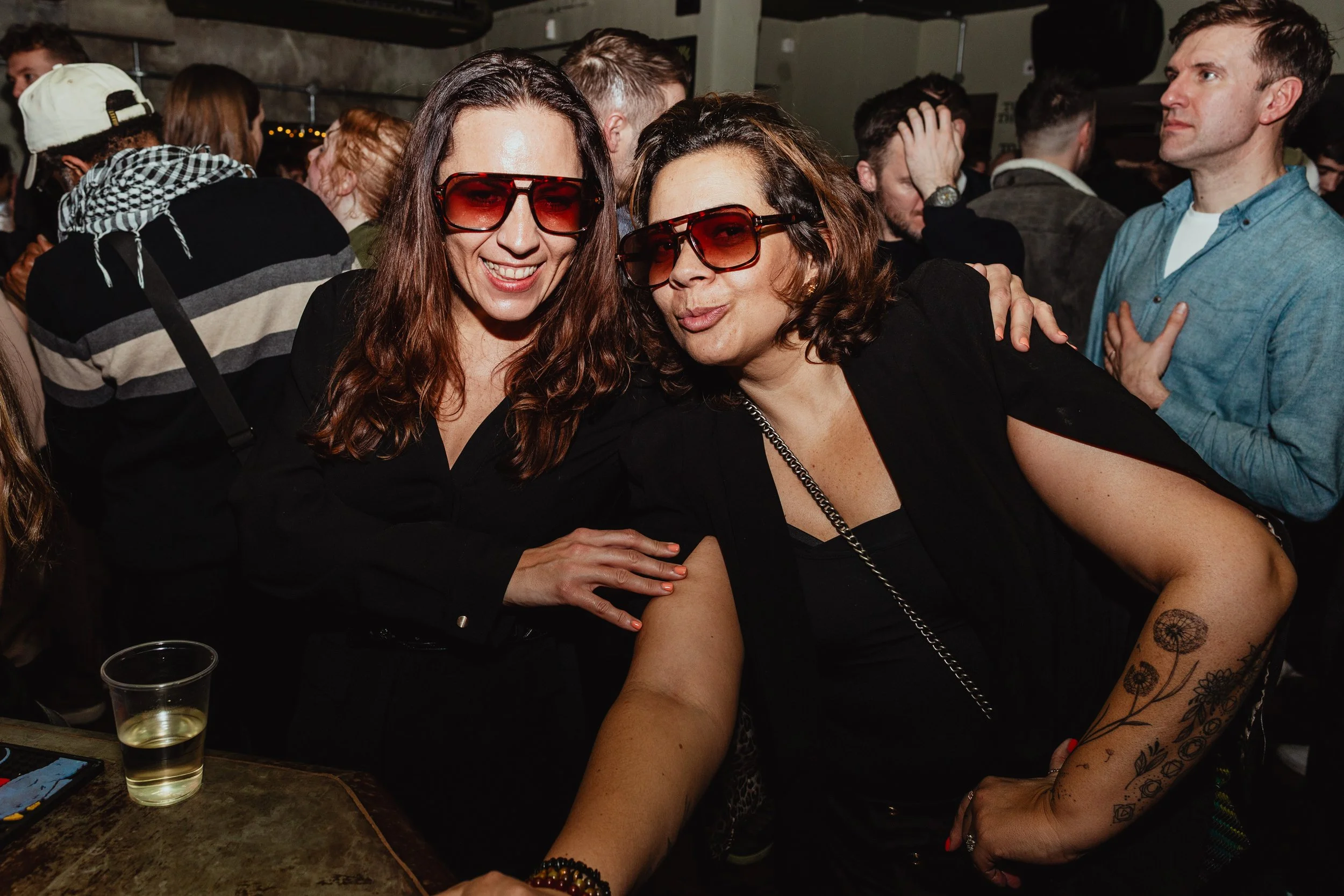 Two women wearing sunglasses are smiling and posing for a photo at a crowded social gathering or party, with people in the background.