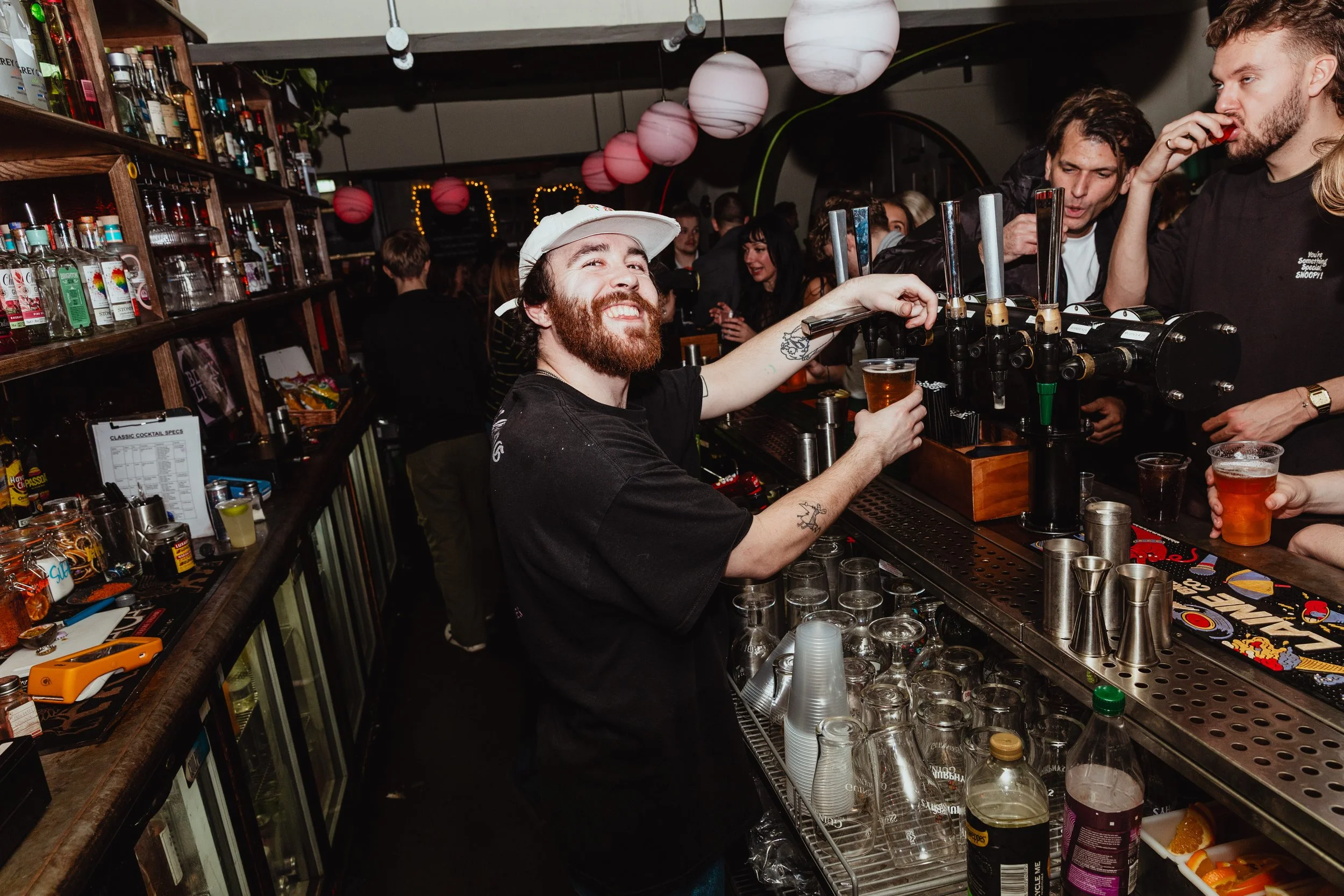 A bartender with a beard and tattoos smiling while pouring a drink at a busy bar. Several customers are in the background enjoying drinks and socializing.
