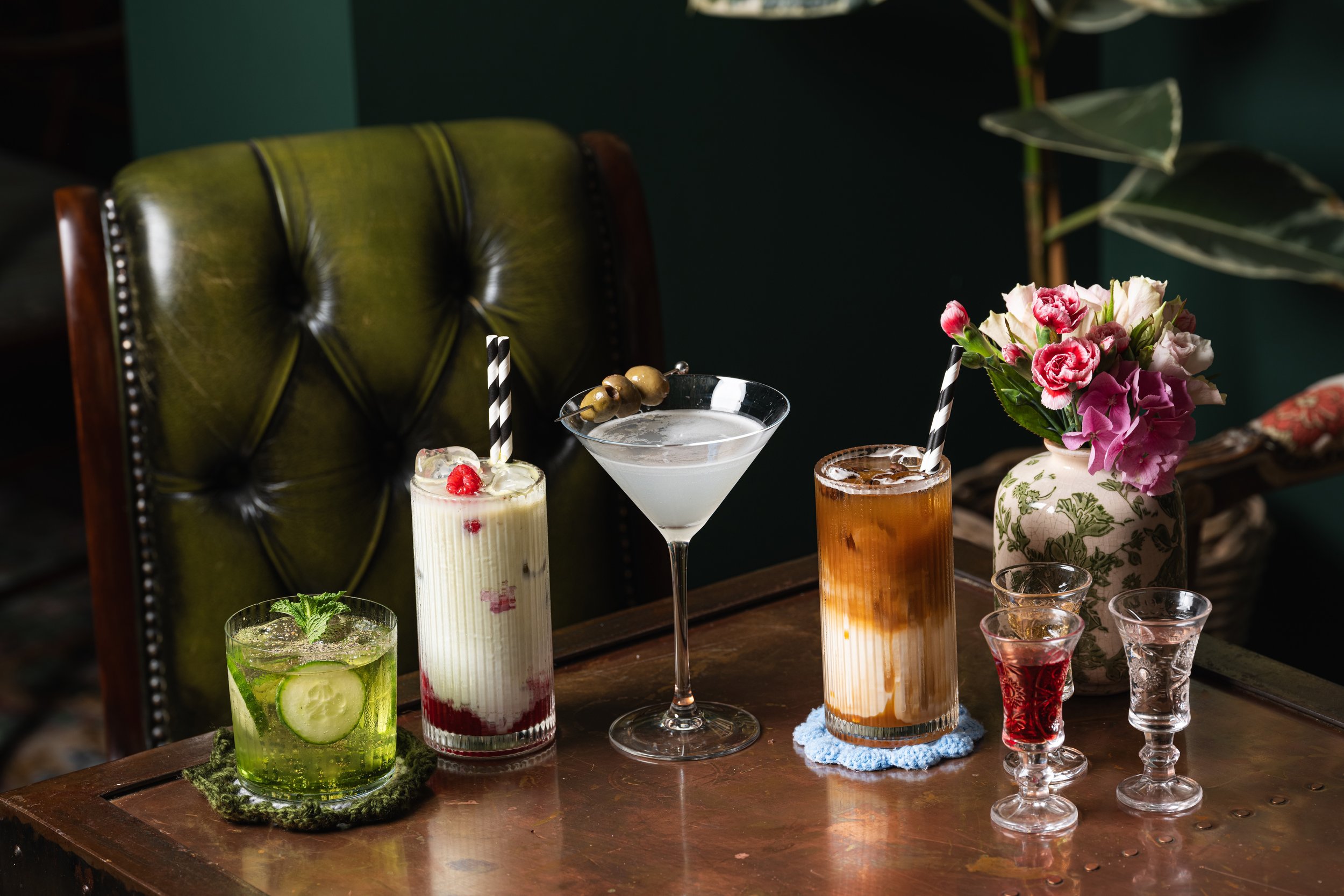 An assortment of six colorful cocktails on a wooden table, with a green leather chair behind them and a vase with pink flowers to the right.