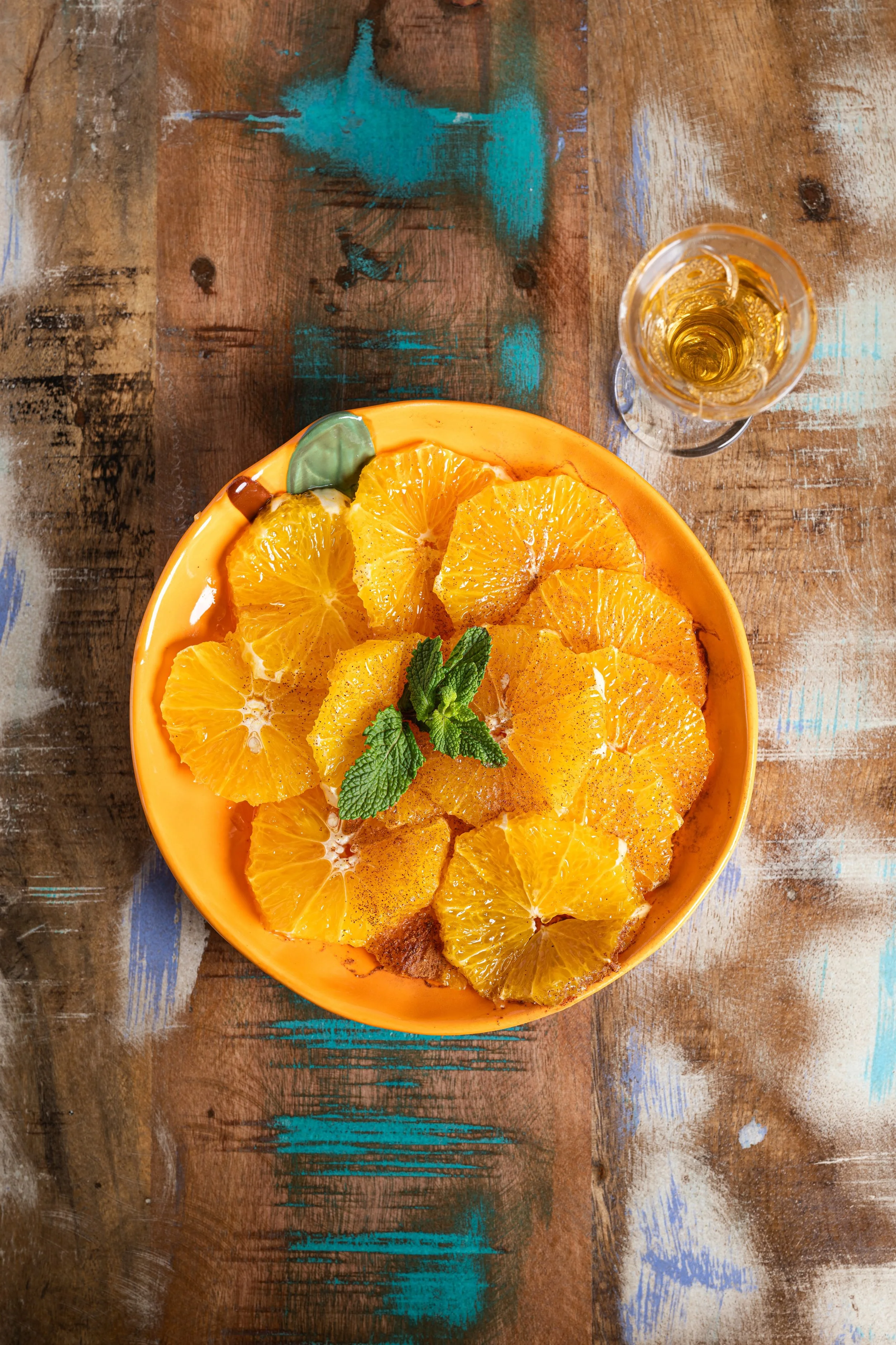 A yellow bowl filled with sliced oranges garnished with mint leaves, placed on a rustic wooden table beside a glass of white wine or champagne.