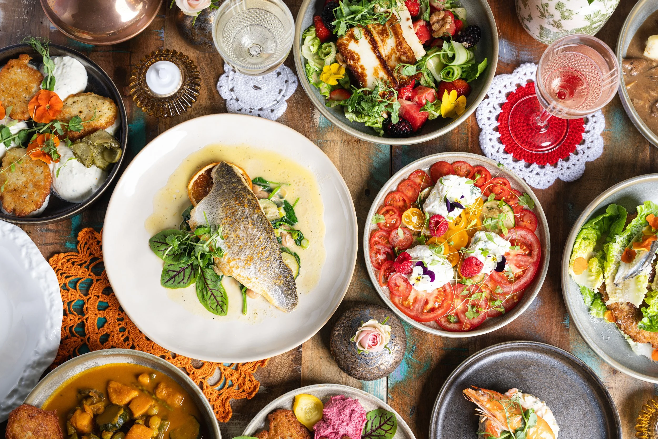 A table with various dishes including salad, fish, tomato and mozzarella, breaded chicken, and a vegetable stew, along with wine glasses and decorative doilies.