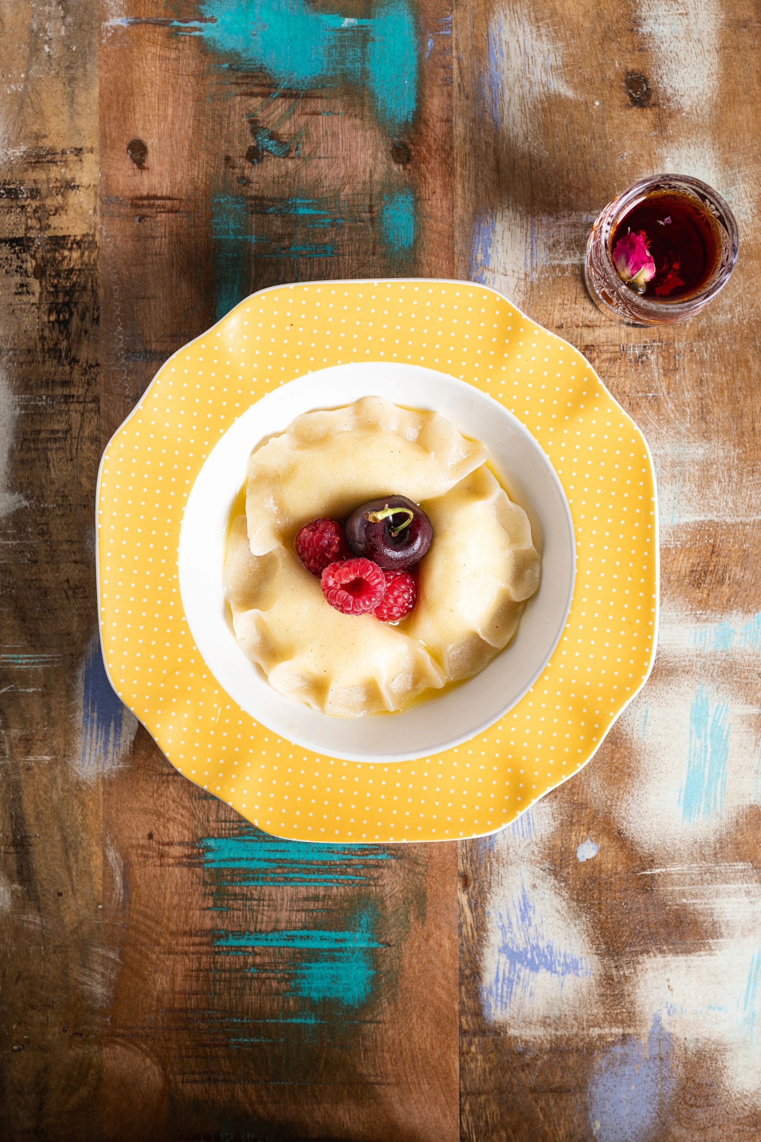 A dessert plate with a piece of yellow custard topped with fresh raspberries and a cherry, on a yellow polka-dot plate, with a glass of tea nearby on a colorful wooden table.