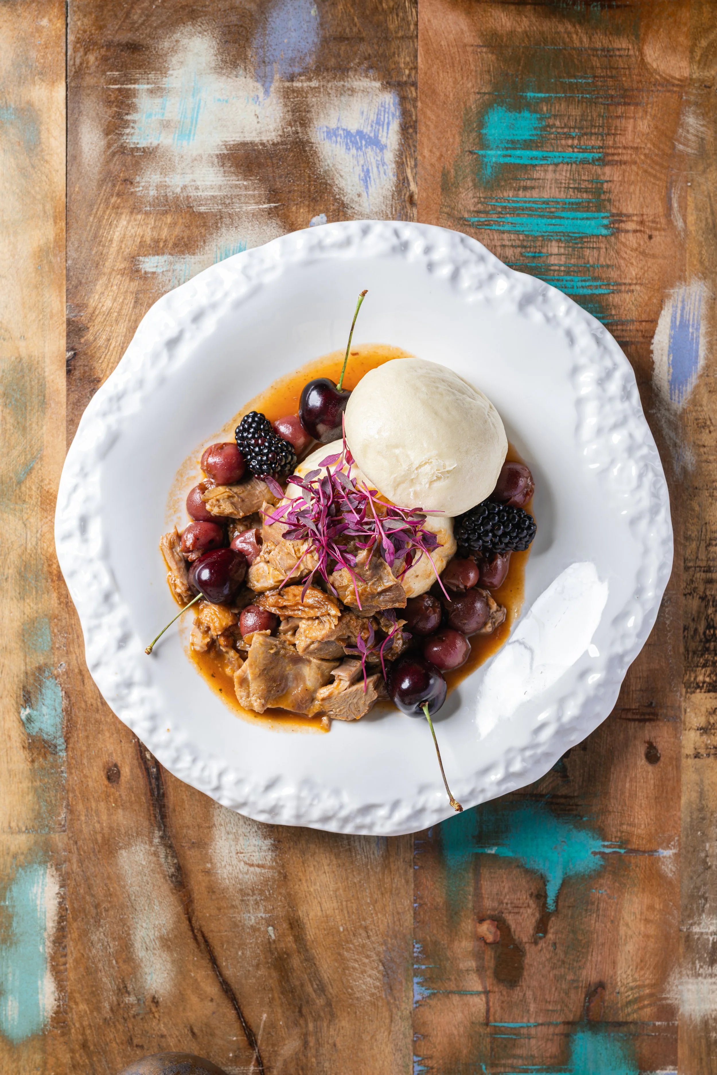 A white decorative plate with meat, grapes, blackberries, a scoop of ice cream, and purple microgreens, on a rustic wooden table with painted details.