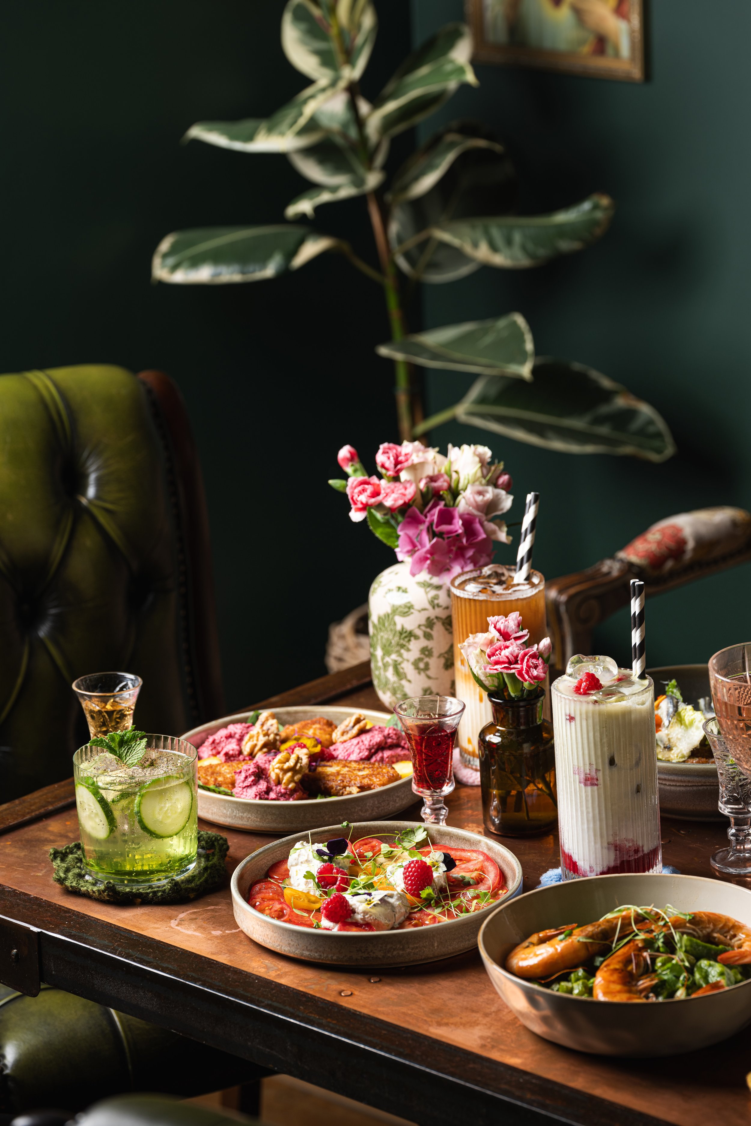 A wooden table with various dishes and drinks, including cocktails, a pizza, and a bowl of shrimp with greens, decorated with flowers and a large houseplant in the background.