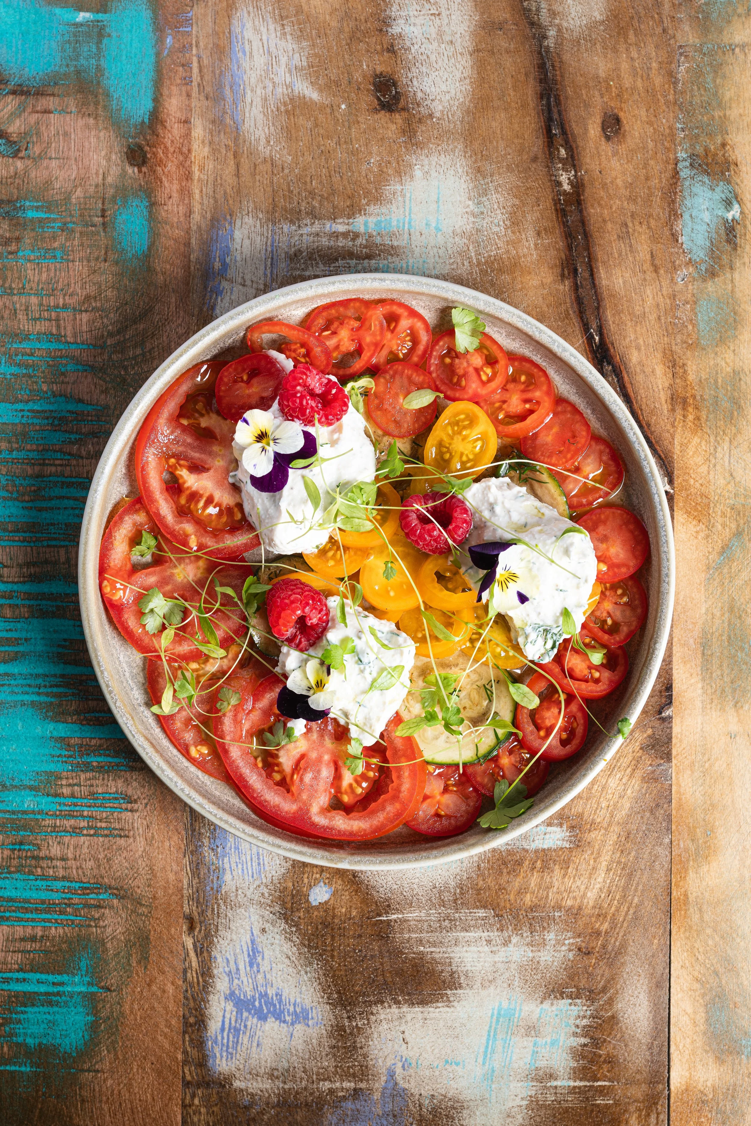 A bowl of sliced tomatoes topped with dollops of cheese or cream, fresh raspberries, small edible flowers, and microgreens, on a rustic wooden table.