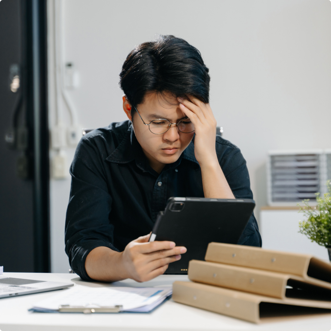 Young man with glasses, sitting at a cluttered desk, looking at a tablet with a confused or stressed expression, holding his forehead.