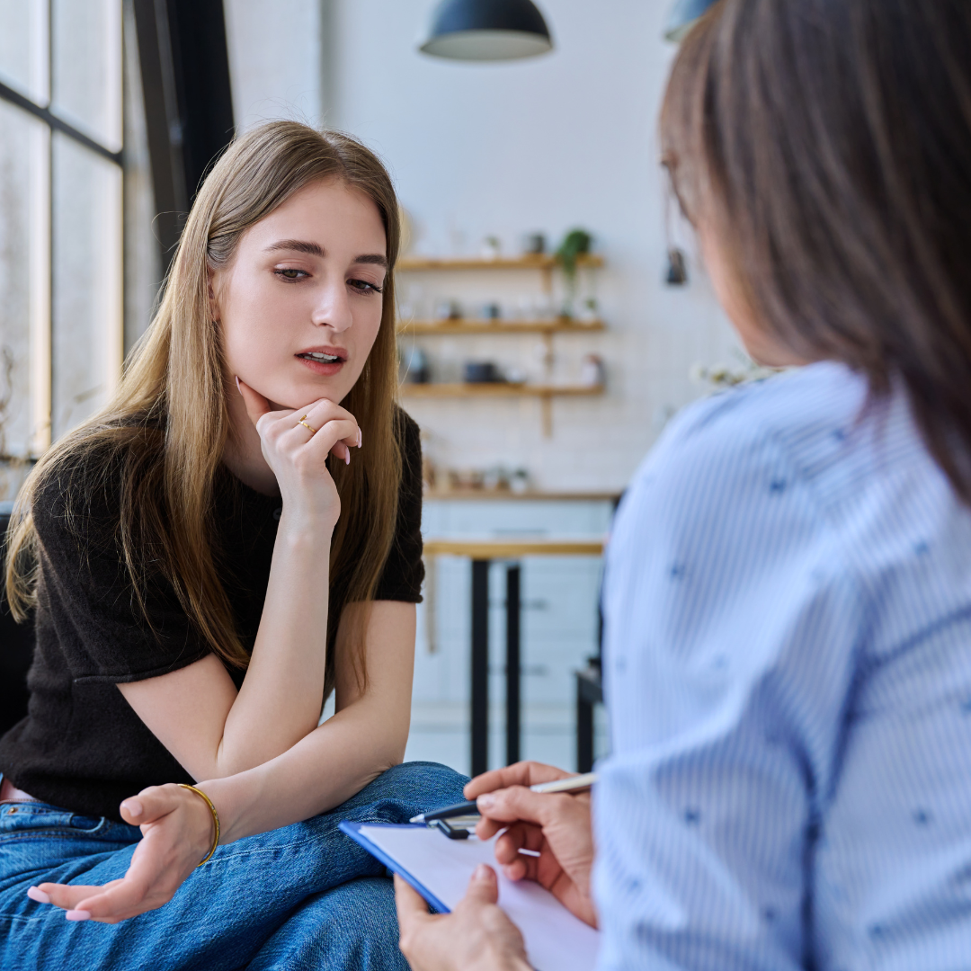 A young woman with long hair is sitting in a counseling session with a therapist, appearing thoughtful as she listens and talks during the discussion.