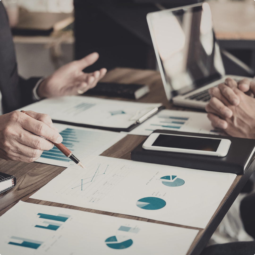 Business meeting with two people discussing financial charts and graphs, with documents, a tablet, a smartphone, and a laptop on a wooden table.