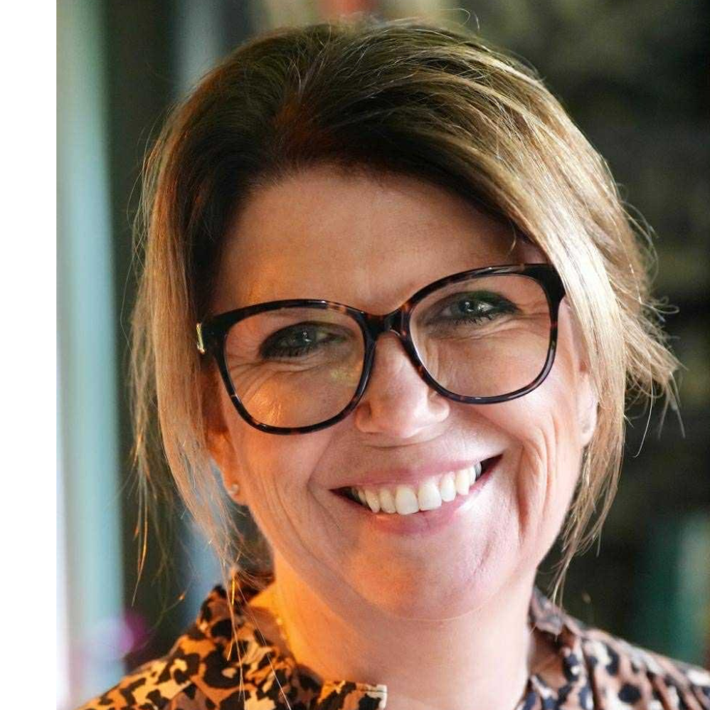 Close-up of a woman wearing black glasses, smiling, with short brown hair and a patterned shirt.