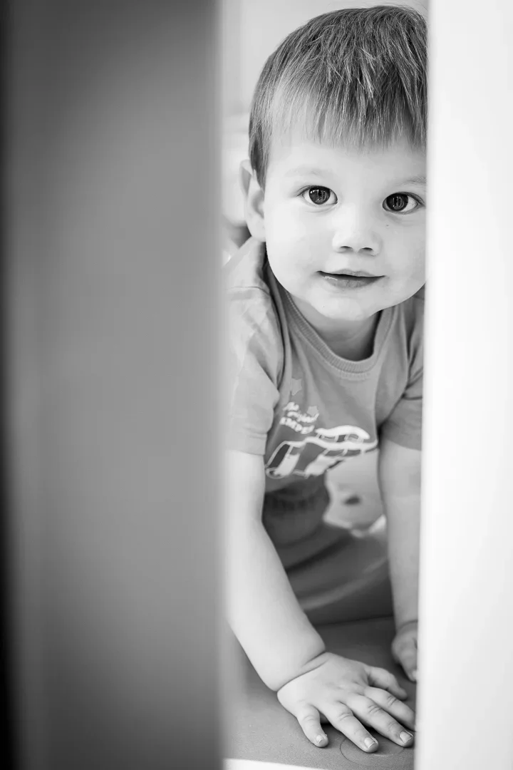 Zwarte en witte foto van een jonge jongen die tussen twee meubels door kijkt, met een nieuwsgierige blik.