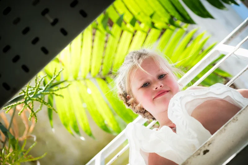 Een jonge meisje met blond haar dat in een witte jurk op een balkon ligt met groene planten en een geperforeerde metalen balk op de voorgrond.