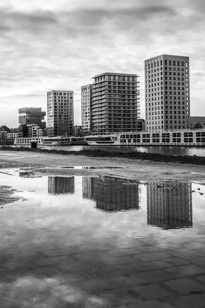 Een zwart-wit foto van een moderne stad met hoge gebouwen langs een rivier of haven, met reflecties in het water.