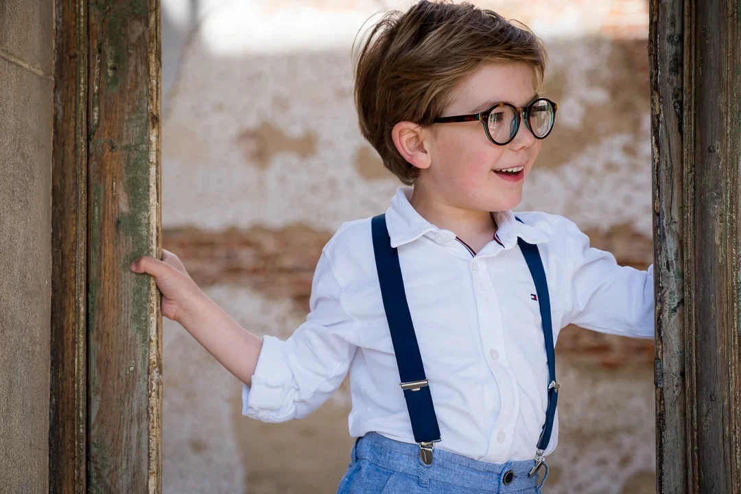 Een jongen met bril, bruin haar, witte blouse, blauwe broek en bretels staat bij een oude, ruwe deuropening en glimlacht.