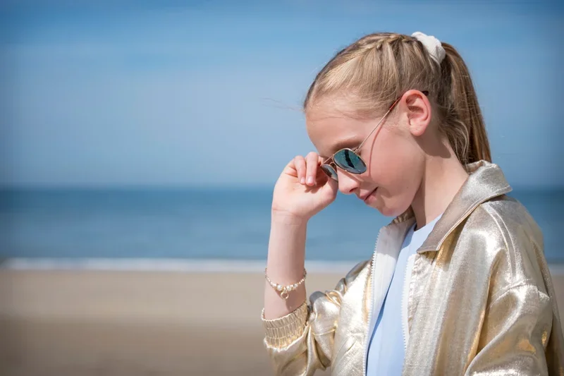 Een meisje met een goudkleurige jas, zonnebril, en een witte haarband op het strand.