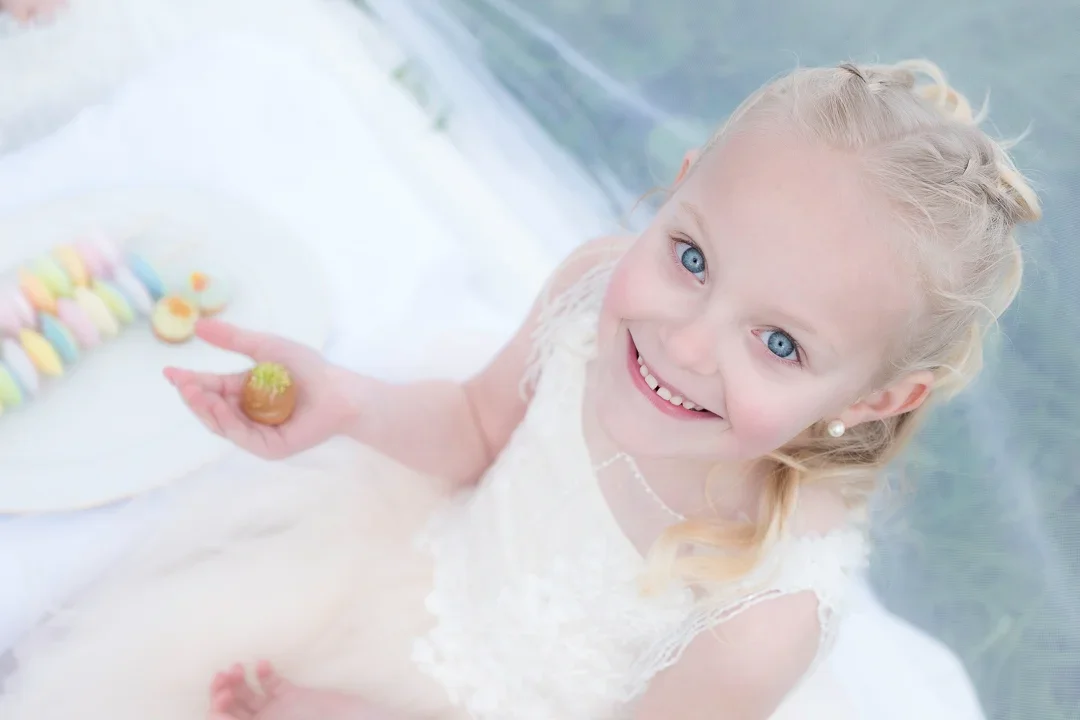 Een lachend meisje met blauwe ogen en blond haar in een witte jurk, zittend in een witte picknick tent met een kleurrijk klein gebakje op haar hand.