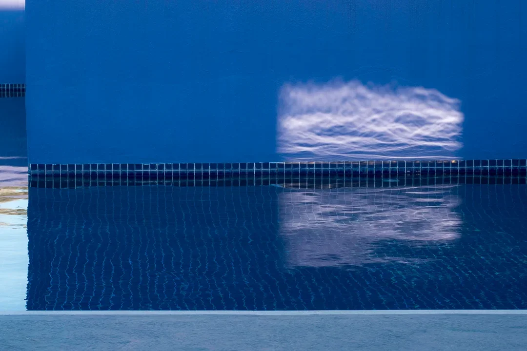 Een zwembad met blauwe tegels en een wateroppervlak die een wolk weerspiegelt, met een blauwe muur op de achtergrond.