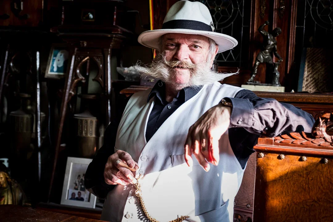 Een oudere man met een grote bos witte haar en snor, lachend, gekleed in een zwarte shirt en een witte vest, draagt een witte hoed met zwarte band, binnen in een antiek interieur.