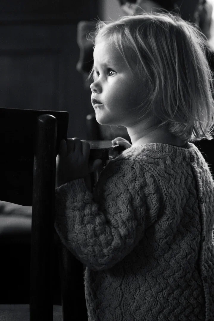 Zwarte en witte foto van jonge meisje met schouders opgetrokken, heldere haar en een gebreide trui, die zich aan een tafel vasthoudt en naar iets kijkt met een serieuze uitdrukking.