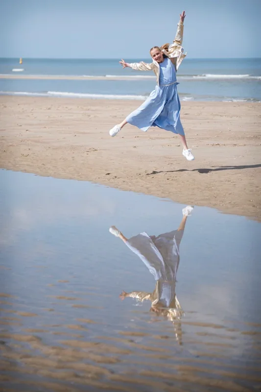 Meisje in blauwe breedvallende jumpsuit en een gouden jas springt op het strand met haar reflectie zichtbaar in een plas water.
