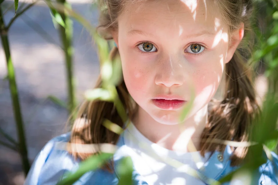 Close-up van een jonge meisje met groene ogen, blonde haar in twee vlechten, omgeven door groene planten, met zonlicht dat door de bladeren schijnt.