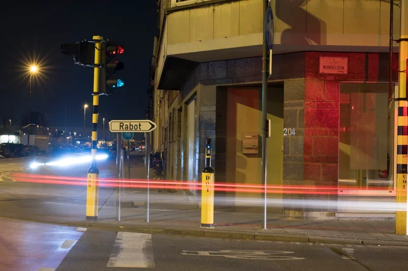 Straathoek met verkeerslichten, een wegwijzer naar Rabot, een gebouw met nummer 204 en straatnaam Wondelgem, lichteffecten van voorbijrazende voertuigen op de weg, 's nachts.