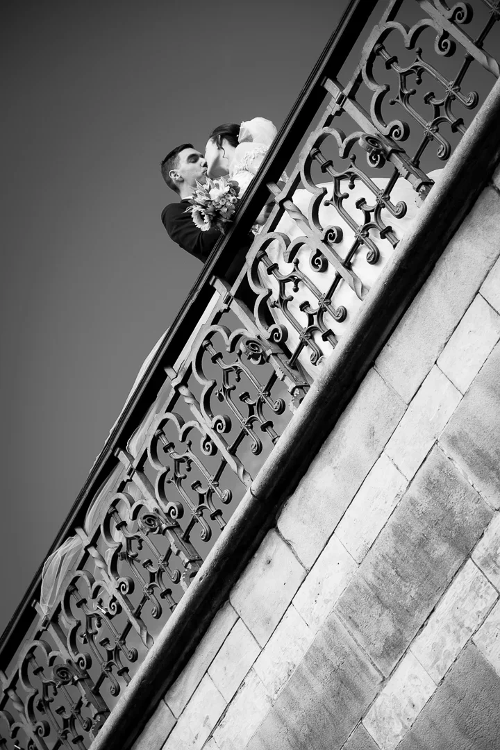 Een zwart-wit foto van een verliefd stel dat op een balkon staat, omgeven door een sierlijk metalen hek. Ze kussen elkaar, de vrouw houdt een bouquet bloemen vast.