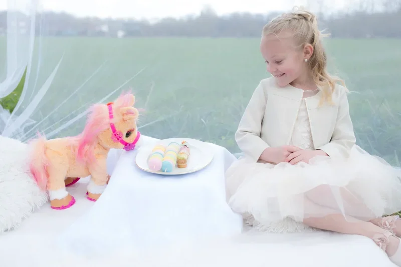 Een lachende jonge meid in een witte jurk en jas zit naast een tafel met een bord met kleurrijke macarons en snoep, en een pluchen speelgoedpaardje met een roze manen en zadel in een witte picknicktent met een groen landschap op de achtergrond.